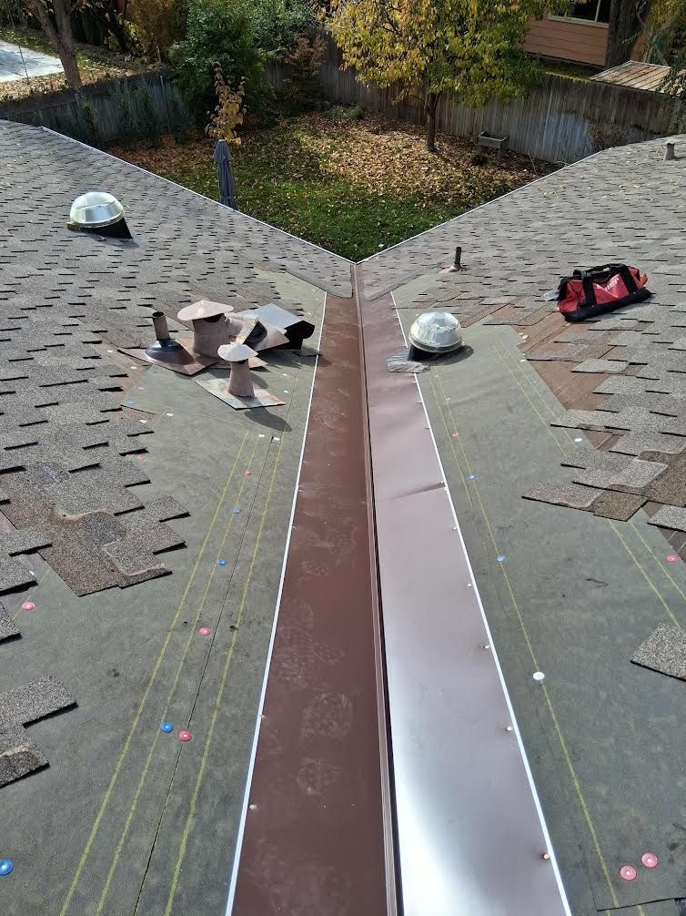A steep residential roof valley with installed brown metal flashing, shingles, vents, and a red tool bag nearby.