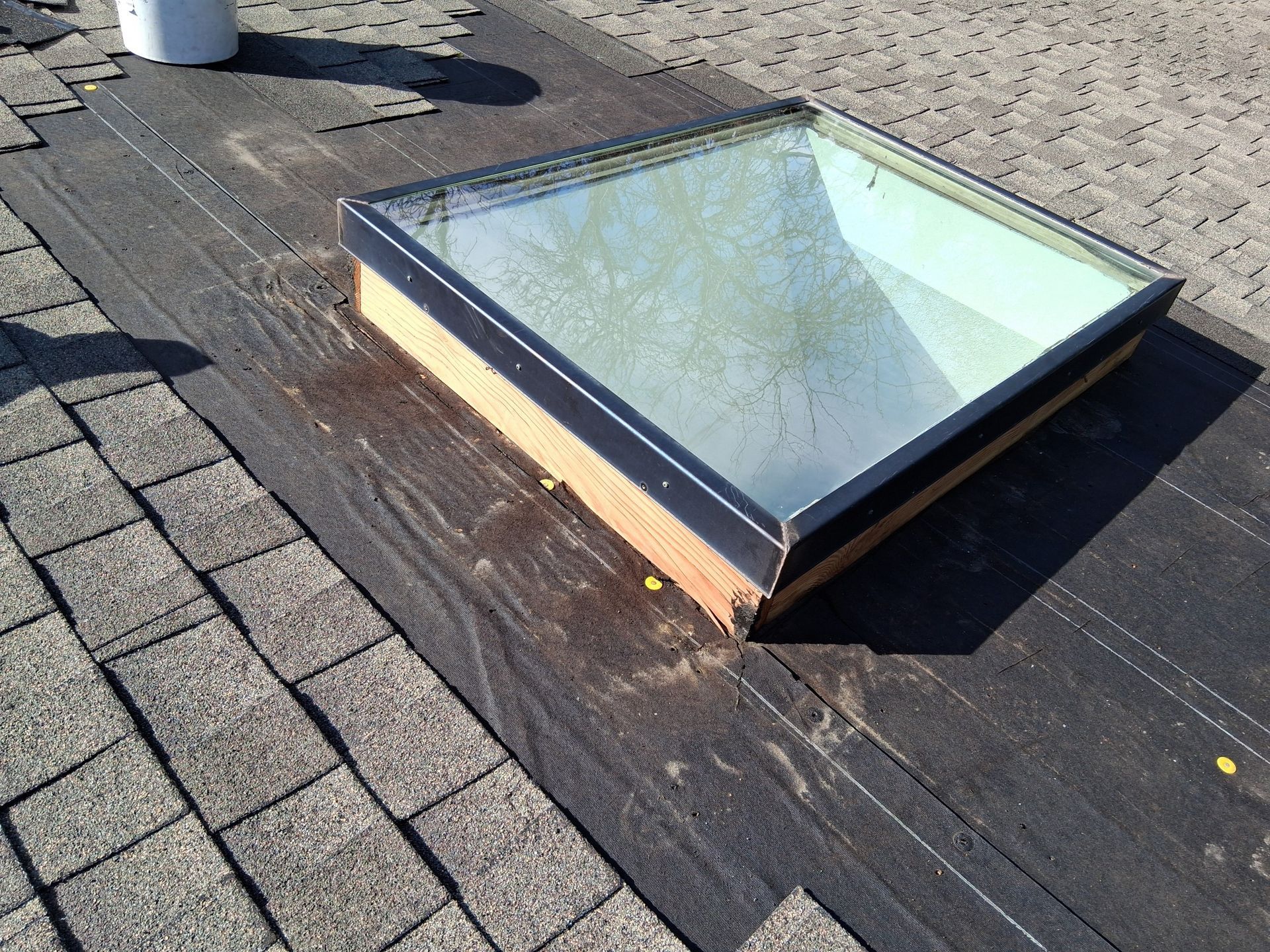 A square skylight sits on a section of a roof with exposed black underlayment next to finished gray roof shingles.
