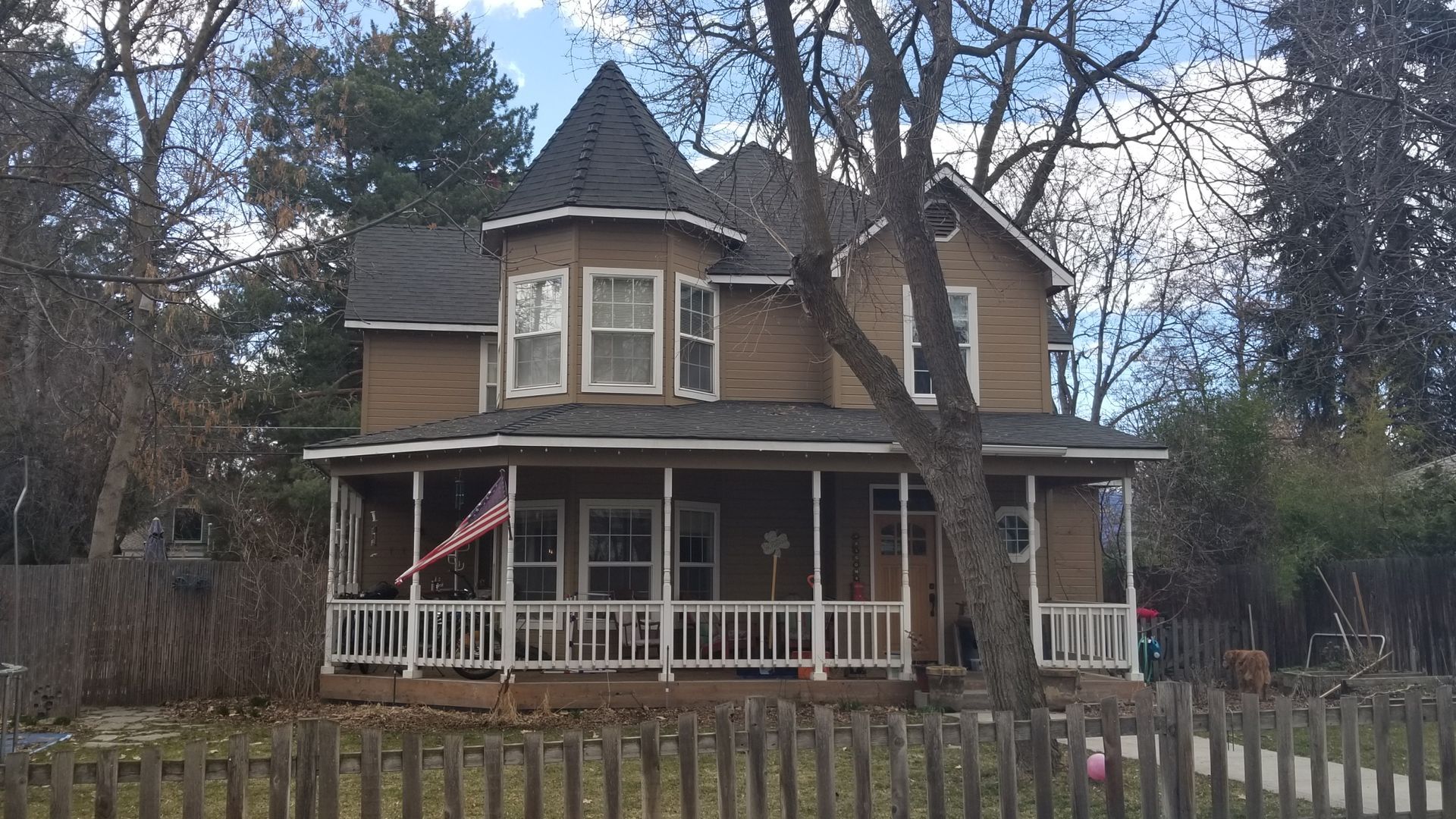 A two-story, tan-colored Victorian-style house with a turret, covered front porch, and a wooden picket fence.