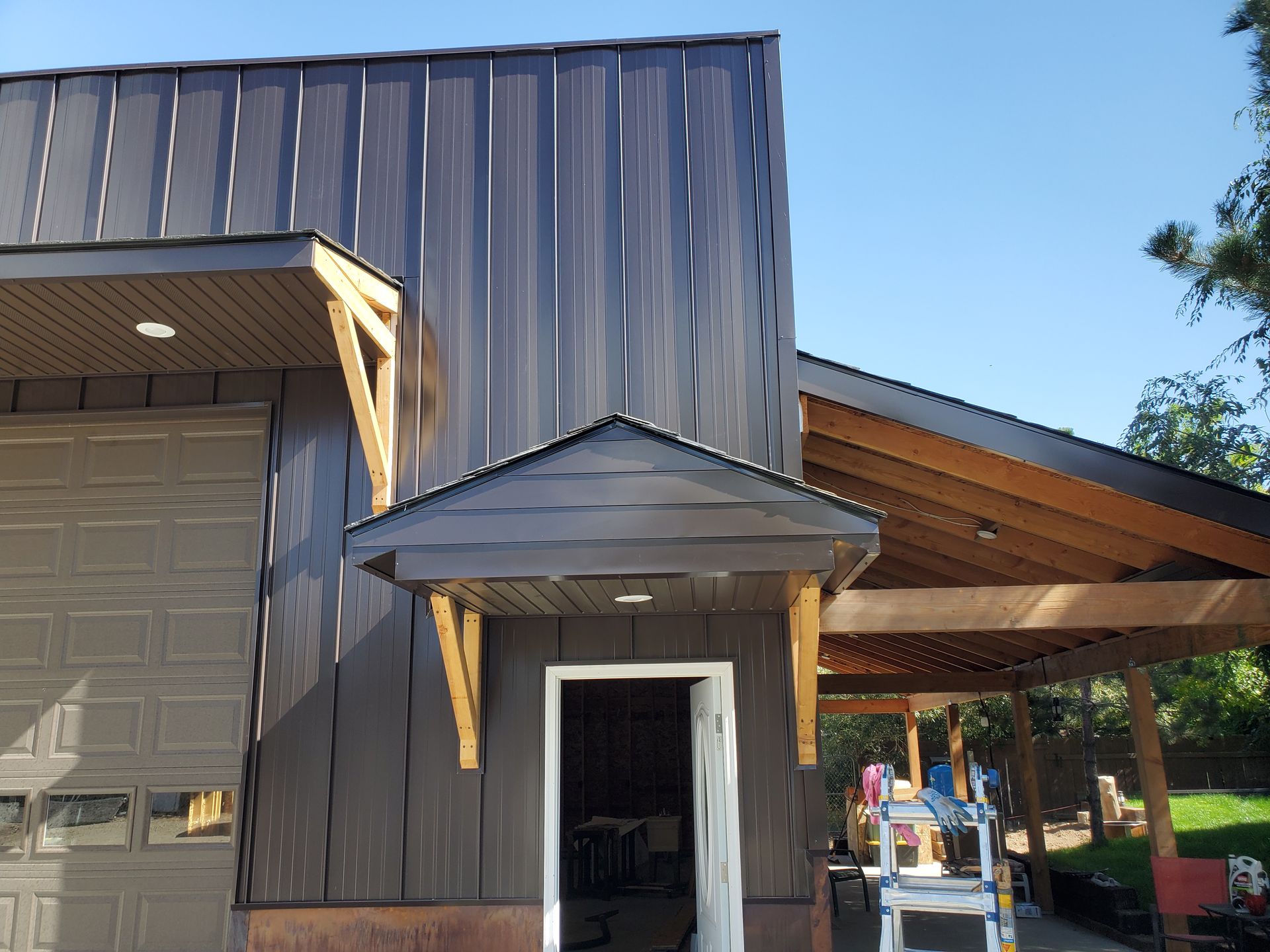A modern building exterior featuring dark vertical metal siding, a garage door, a small entry porch, and a wooden patio.