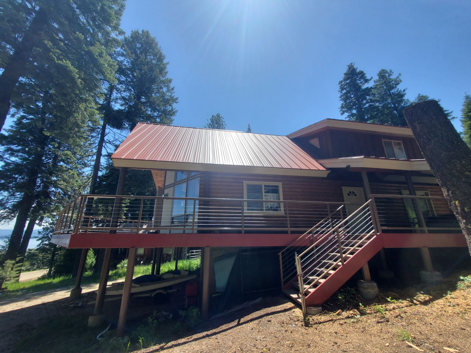 A wooden cabin with a red metal roof and deck, surrounded by tall pine trees on a sunny day.