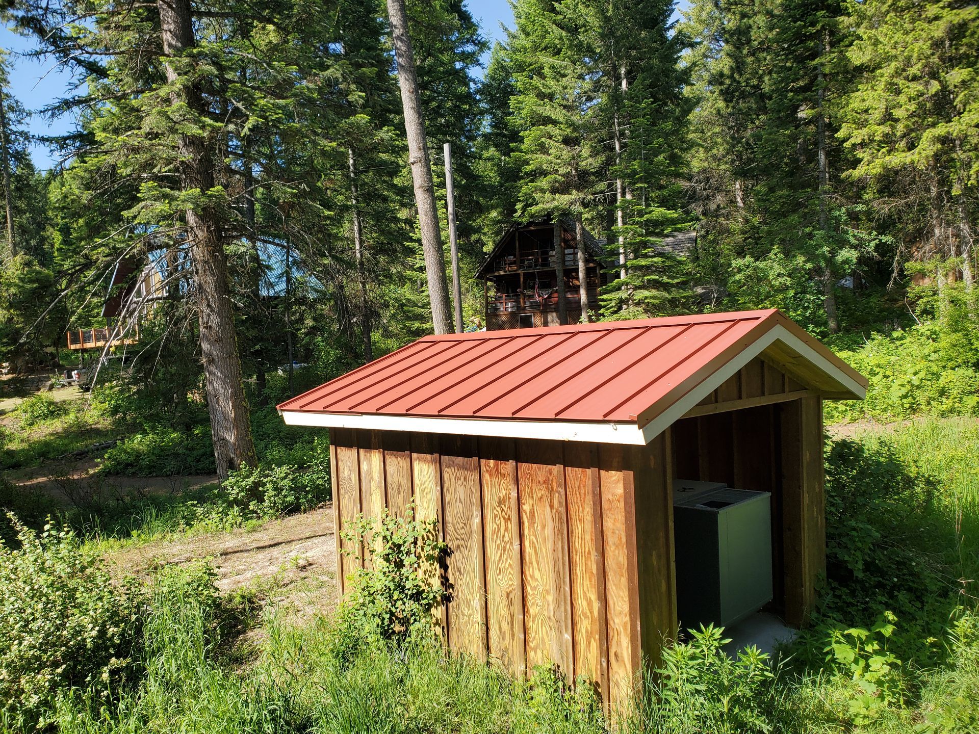A small wooden shed with a bright red metal roof stands in a sunny, forested area with a log cabin in the background.
