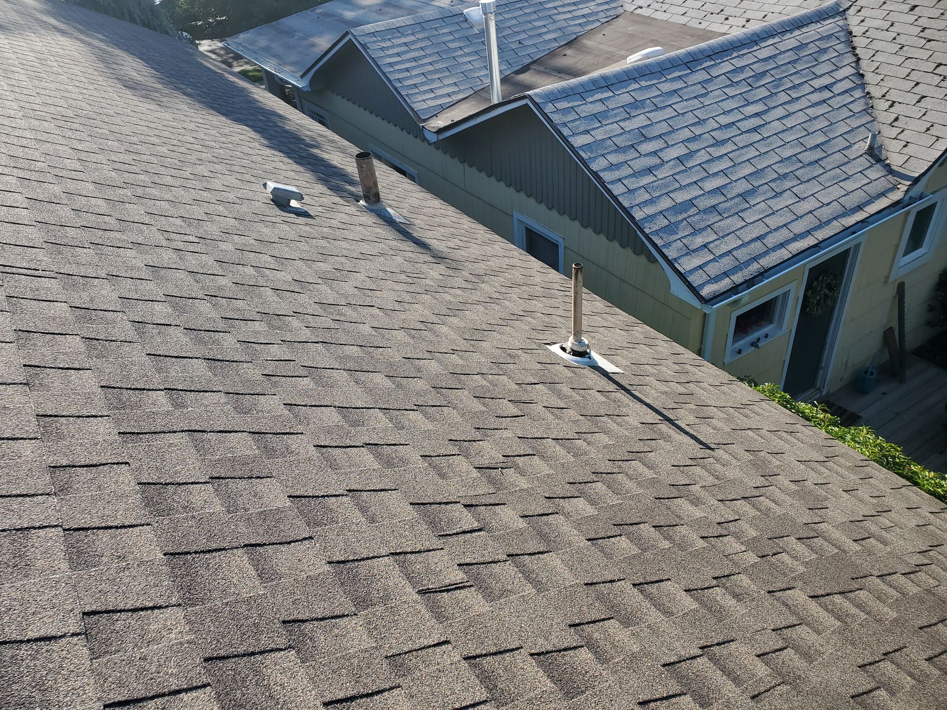 A high-angle view of a gray asphalt shingle roof with several ventilation pipes and an adjacent house section.