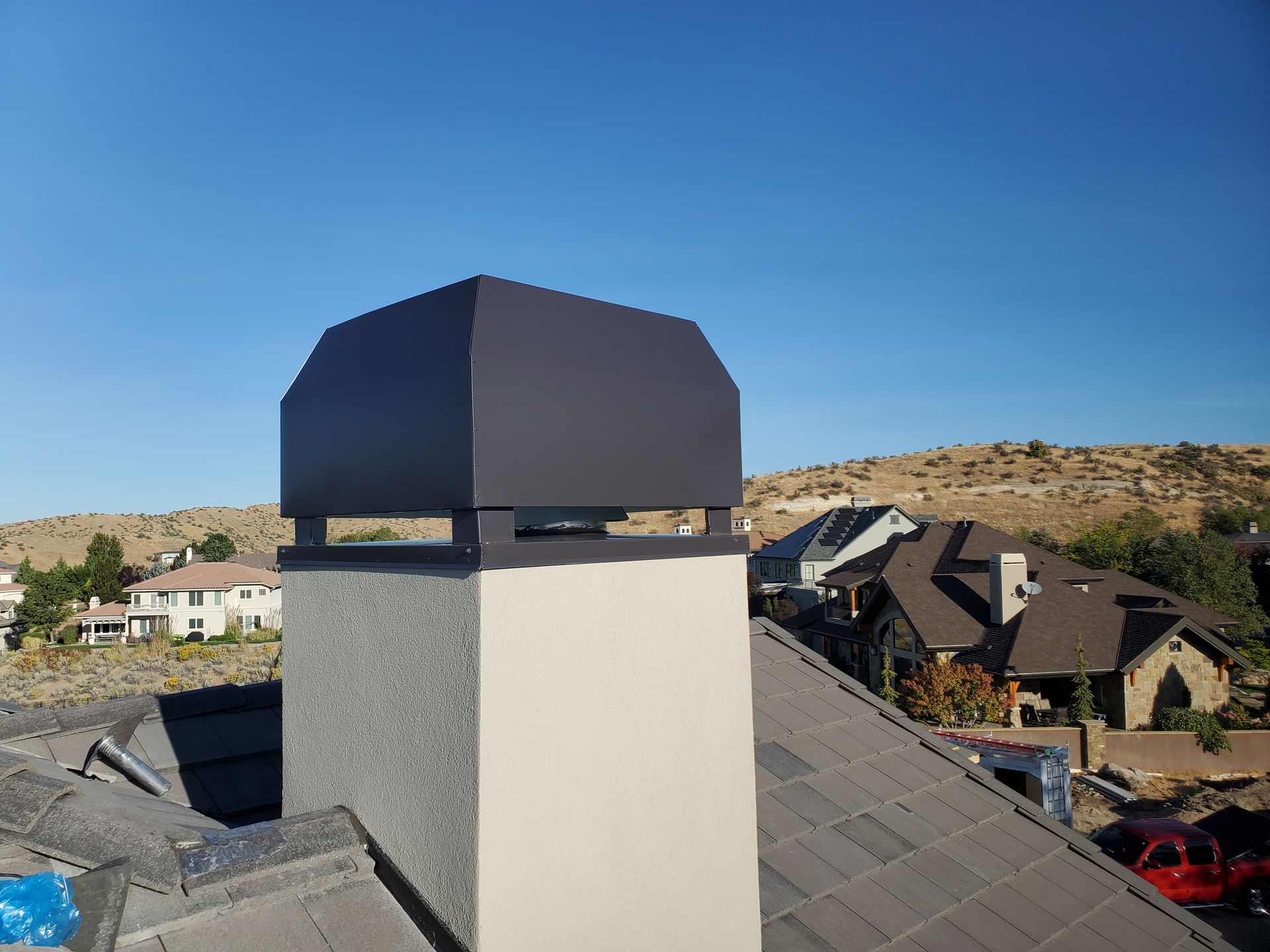A square tan stucco chimney topped with a large dark gray metal cap, viewed from a residential roof on a sunny day.
