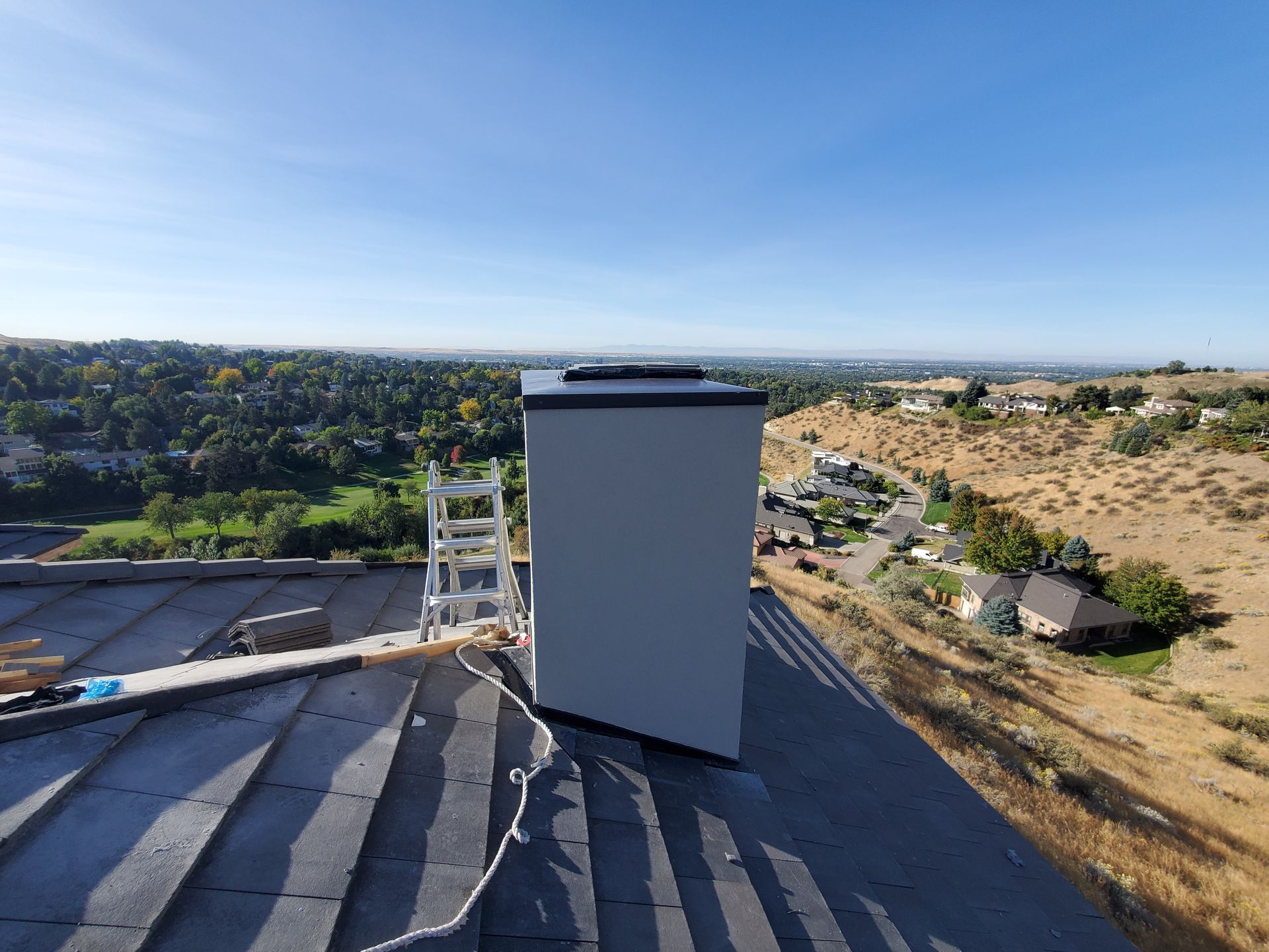 A tall grey chimney structure sits on a residential rooftop, with a ladder leaned against it overlooking a distant valley.