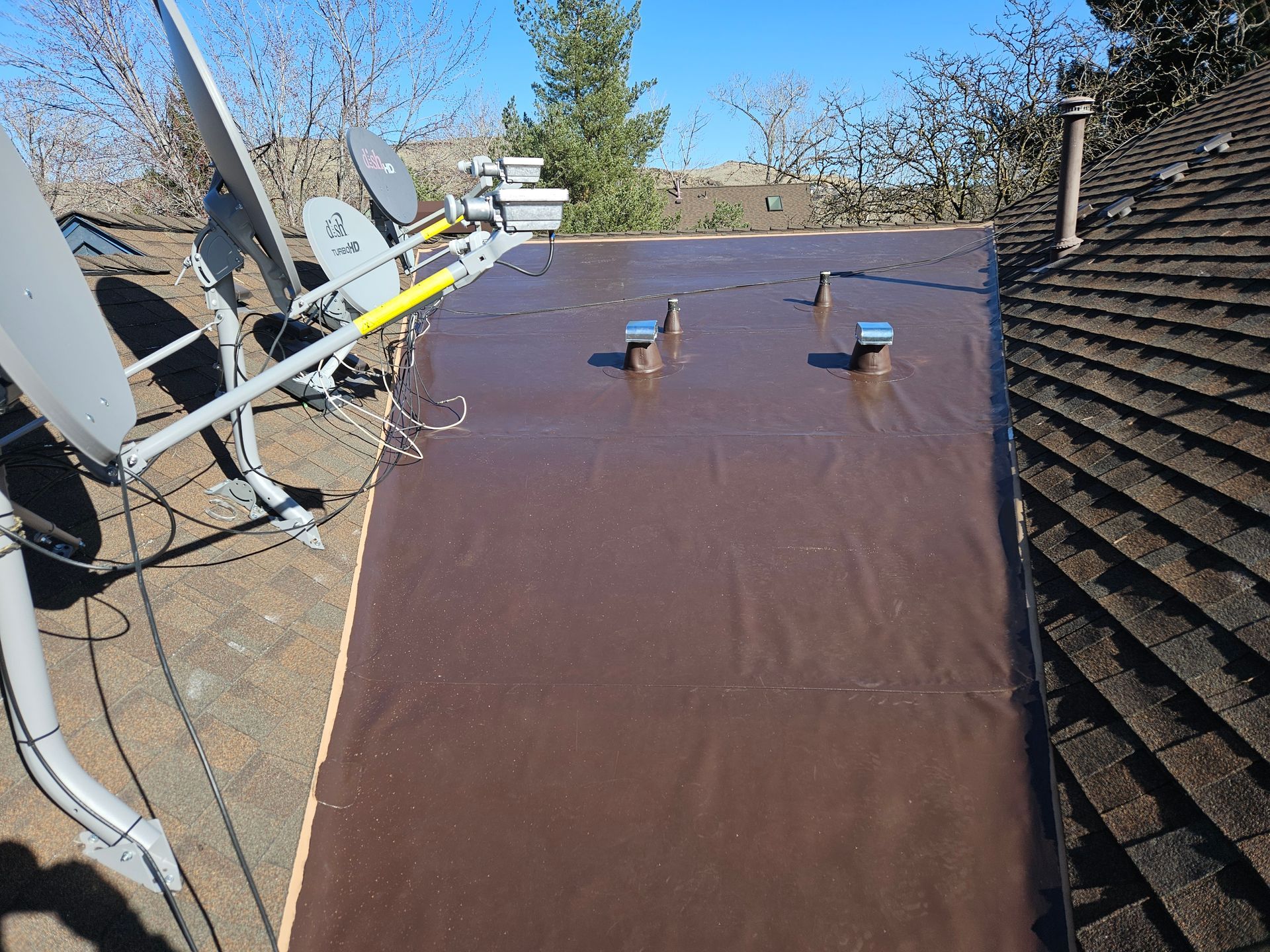 A flat brown roofing section with four vents installed on a residential roof next to shingled areas and satellite dishes.