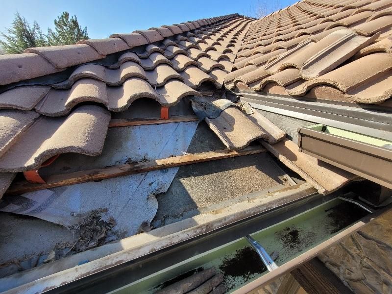 A damaged roof section with missing tiles exposing the underlying wood battens and gray roofing felt above a dirty gutter.