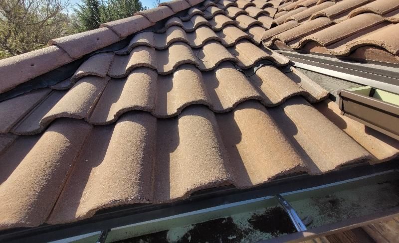 A high-angle view of brown, wavy concrete roof tiles leading to a gutter filled with debris.