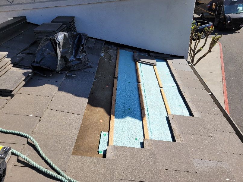 An ongoing roof repair showing exposed wooden beams and light blue insulation panels amidst gray concrete roofing tiles.