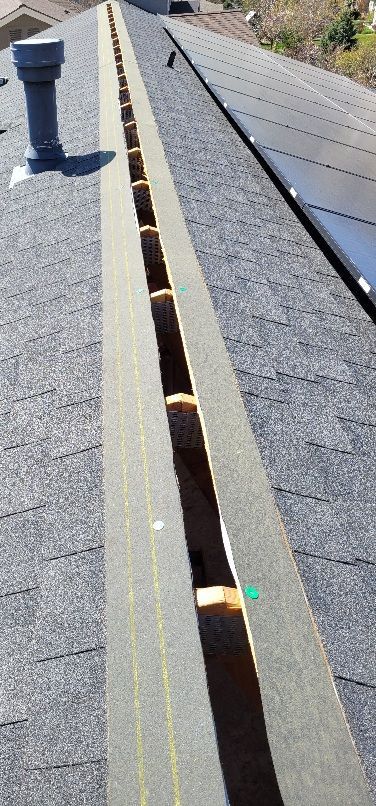 An open ridge vent on a shingled roof, revealing rafters and an interior gap, next to solar panels and a roof vent pipe.