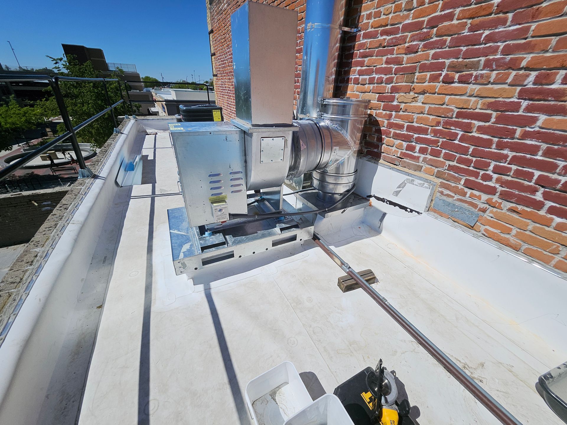A rooftop HVAC unit with silver ductwork stands on a white flat roof next to a brick wall under a clear blue sky.