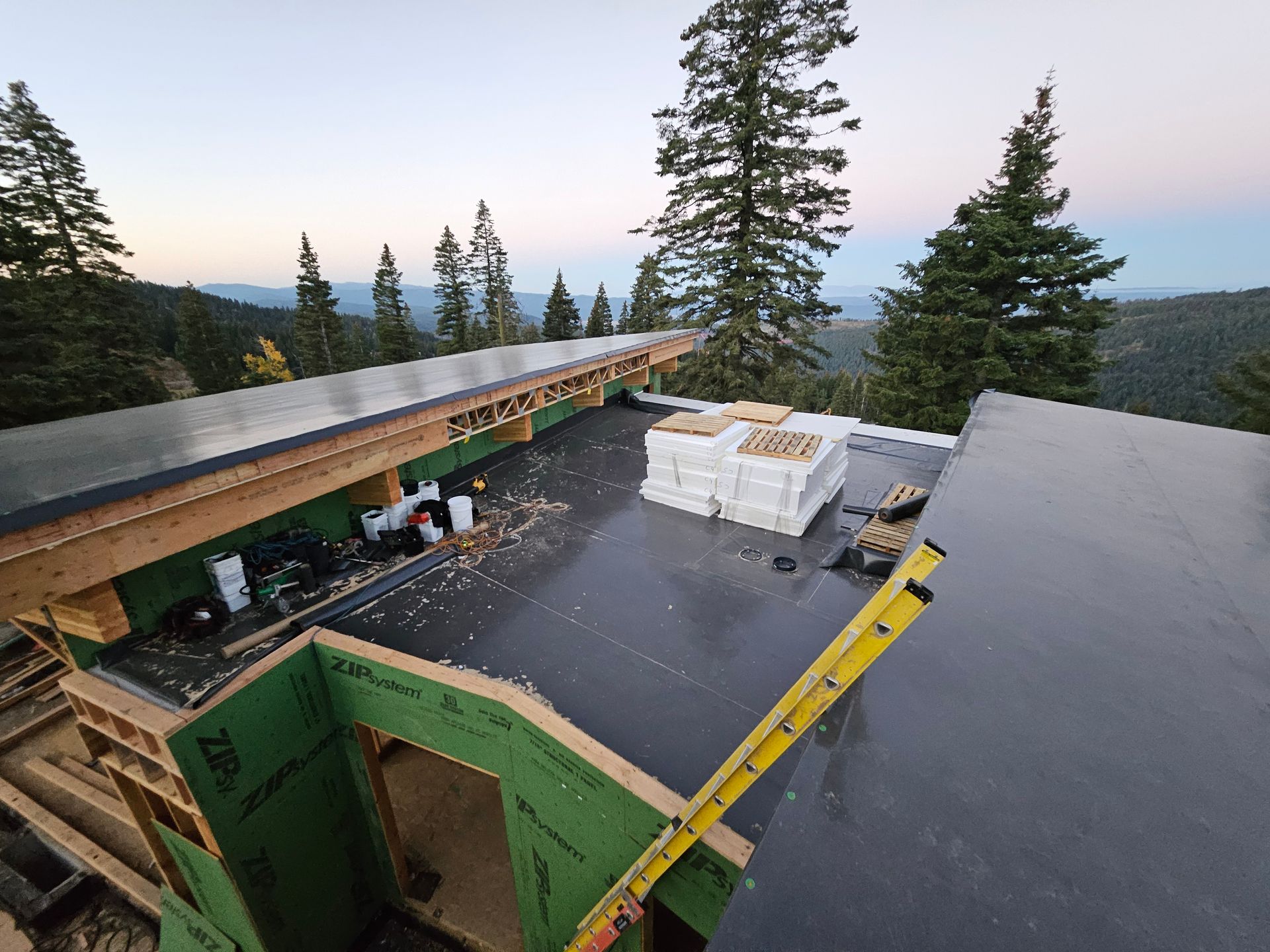 An aerial view of a building under construction featuring a flat, dark roof, wooden framing, green wall panels, and a ladder.