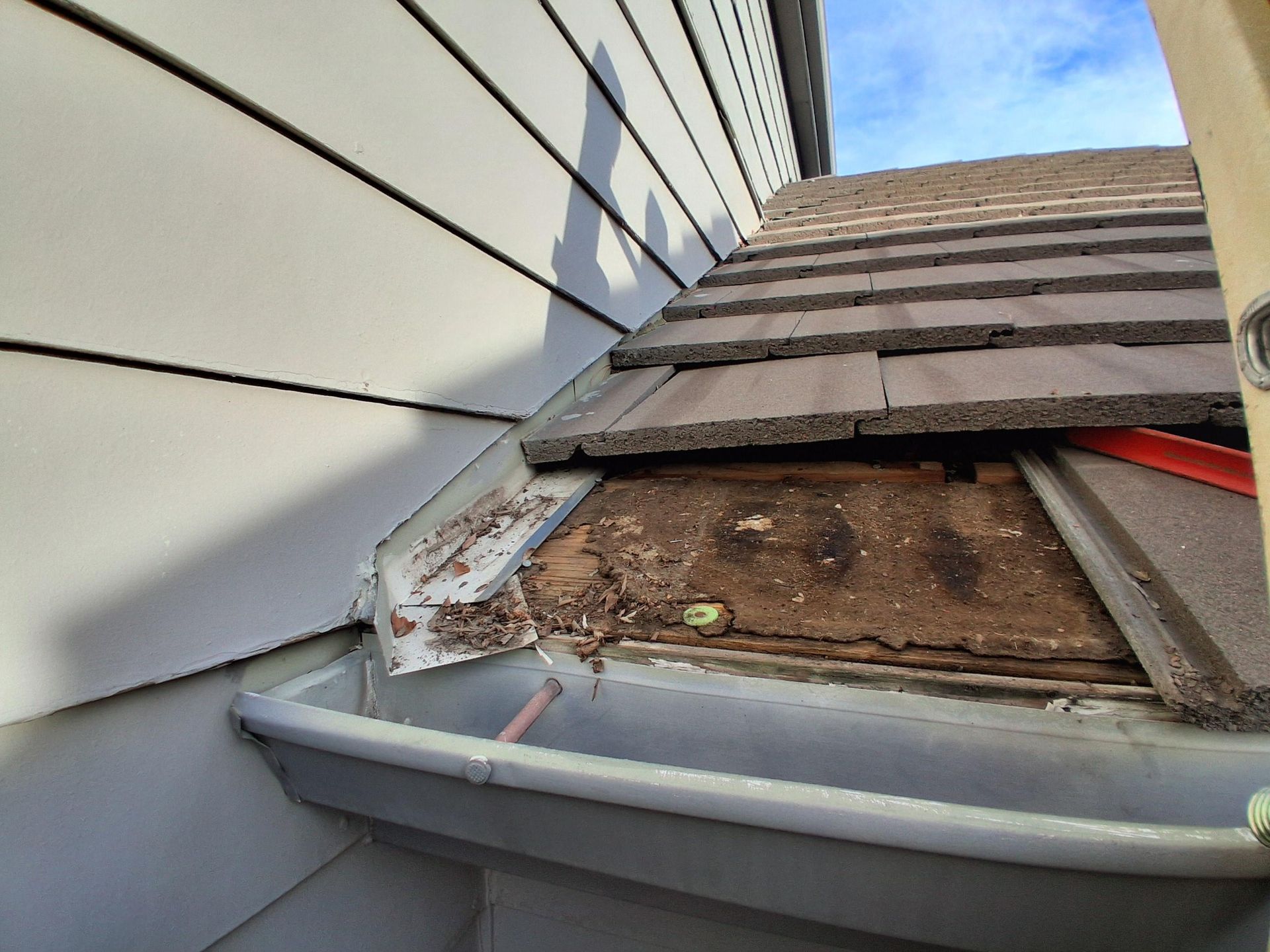 Roof valley with missing tiles, exposed wooden decking, and debris leading into a gutter against beige siding.