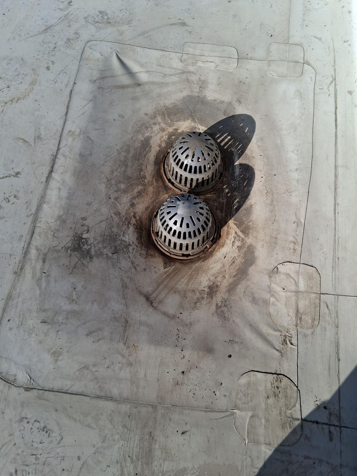 Two metallic dome-shaped roof drains sit side-by-side on a weathered, light-gray flat roof surface.