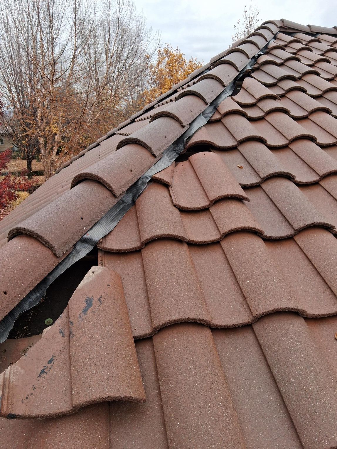 A section of a damaged, brown tiled roof with a broken tile and a gap along the ridgeline.