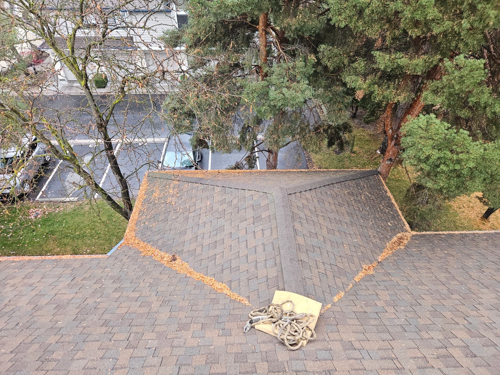 A view from above looking down a dark gray shingled roof with a pile of rope at the base, near trees and a parking lot.
