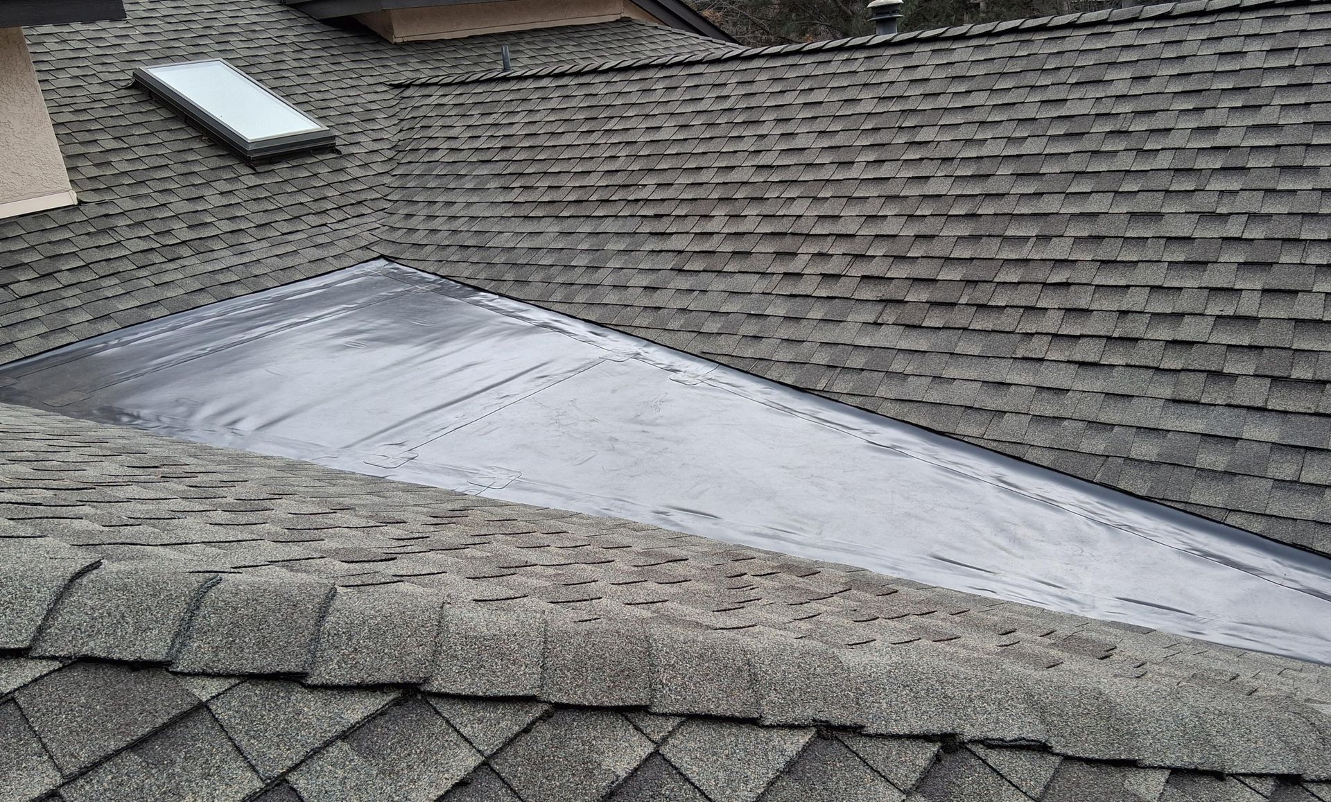 A flat, grey membrane roof section sits in a valley between pitched, grey shingled roof slopes with a skylight nearby.