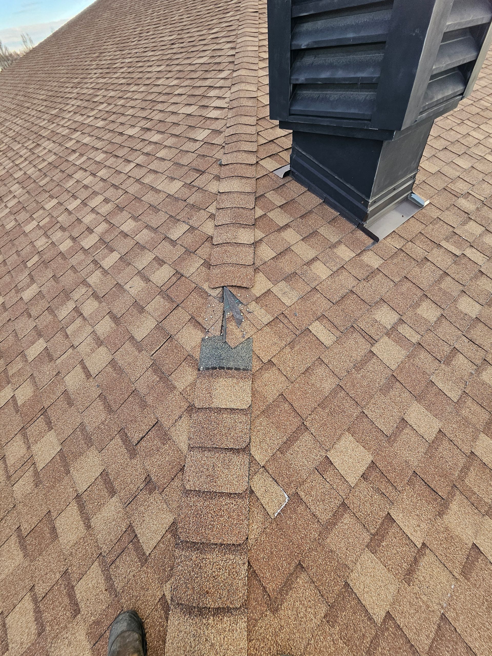 A high-angle view of a brown asphalt shingle roof, showing a section of ridge cap shingles with a repair patch.