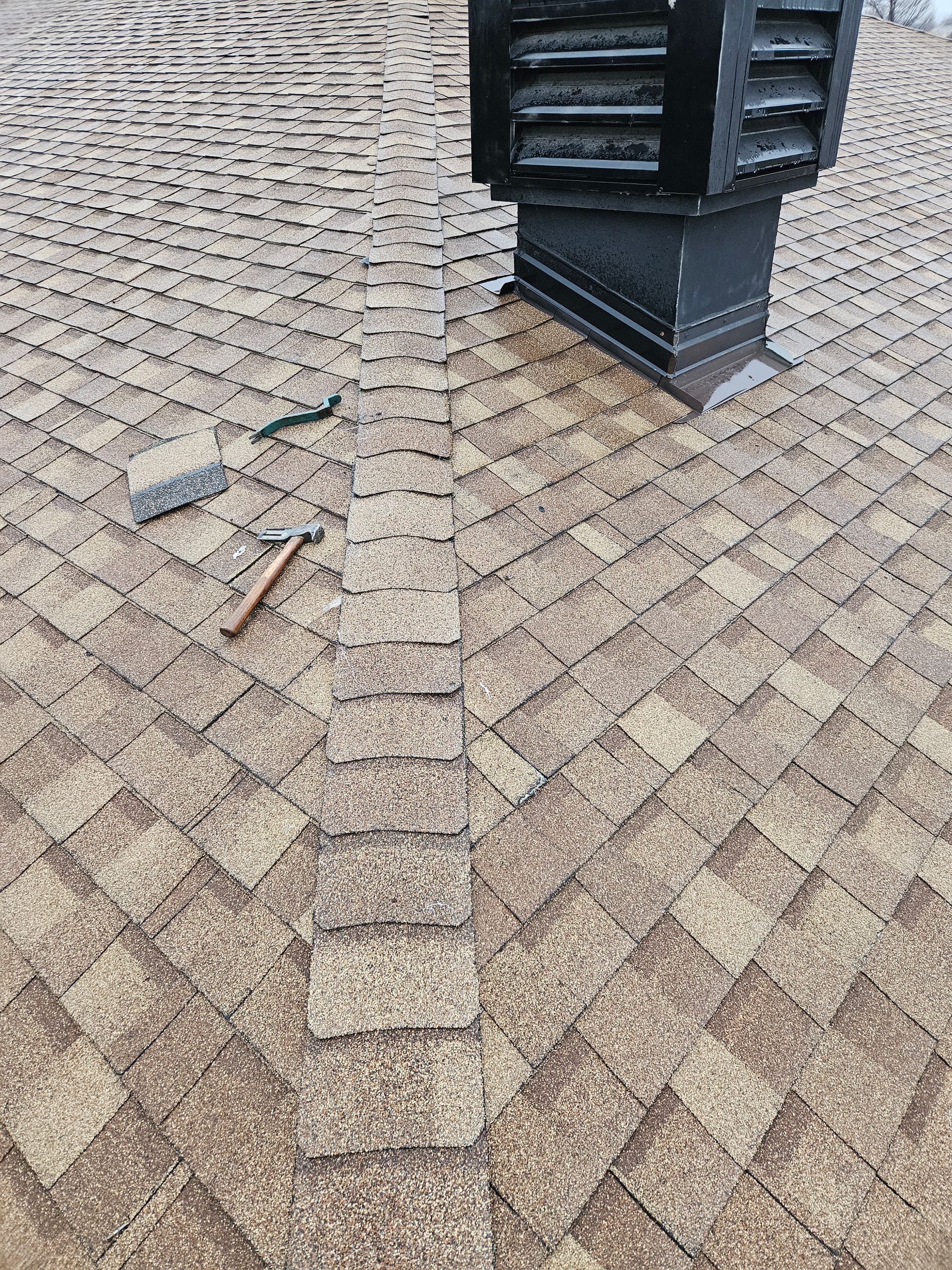 A hammer and block rest on a brown shingled roof next to a chimney and a finished ridge cap installation.