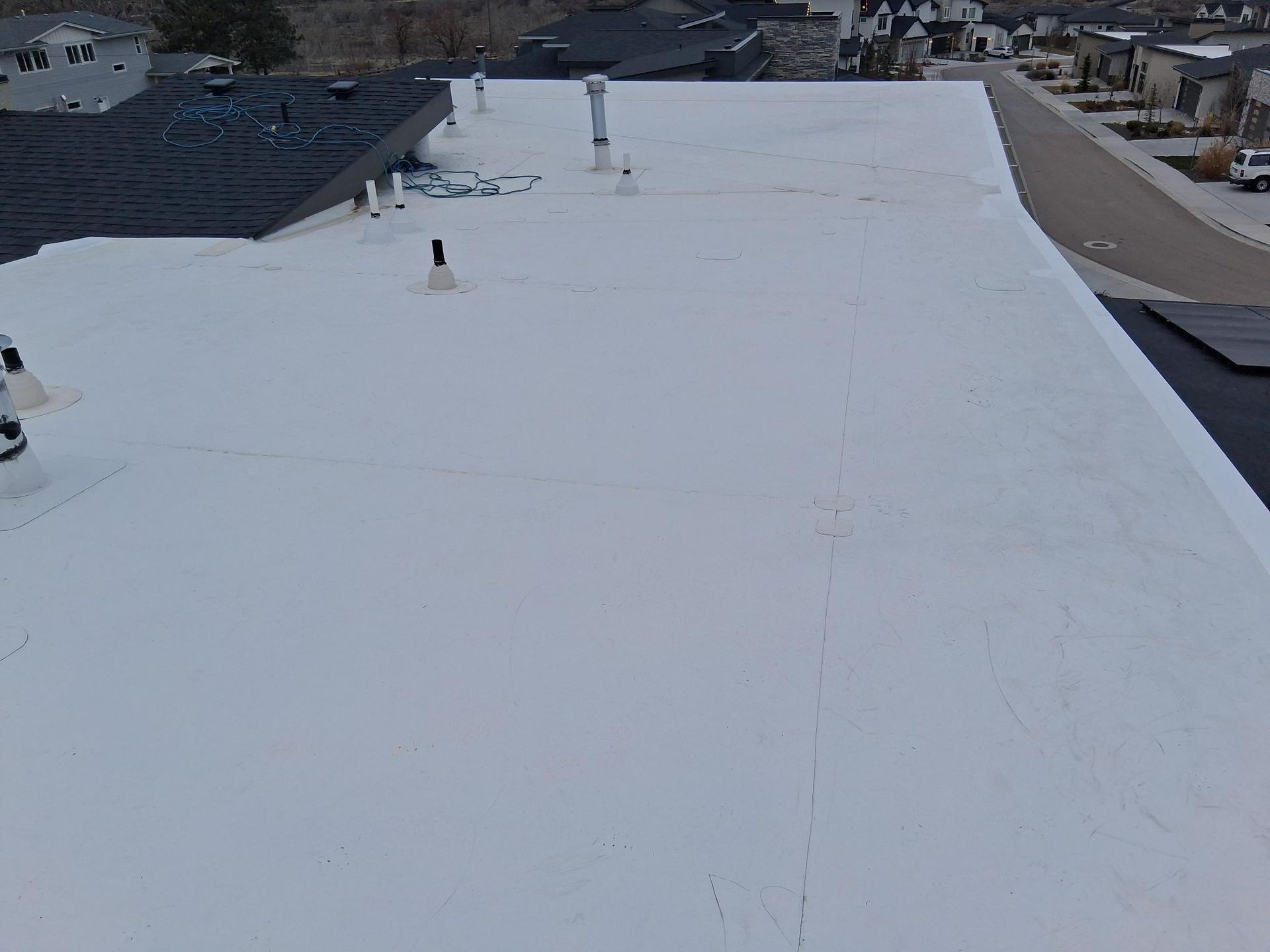 A white, flat roof surface featuring multiple ventilation pipes, viewed from an elevated vantage point above a neighborhood.