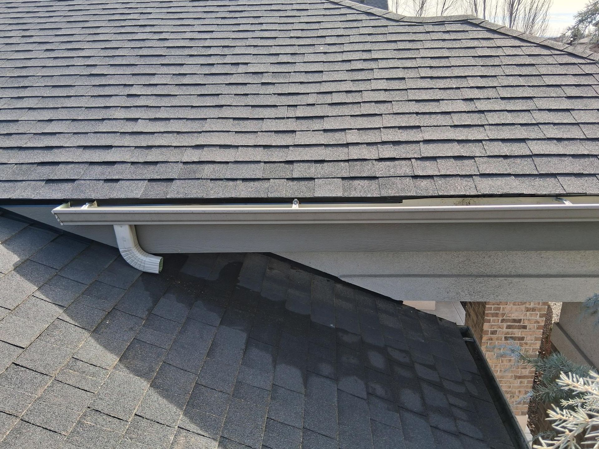 A tan metal gutter attached to the eaves of a shingled roof with a white downspout elbow at the corner.