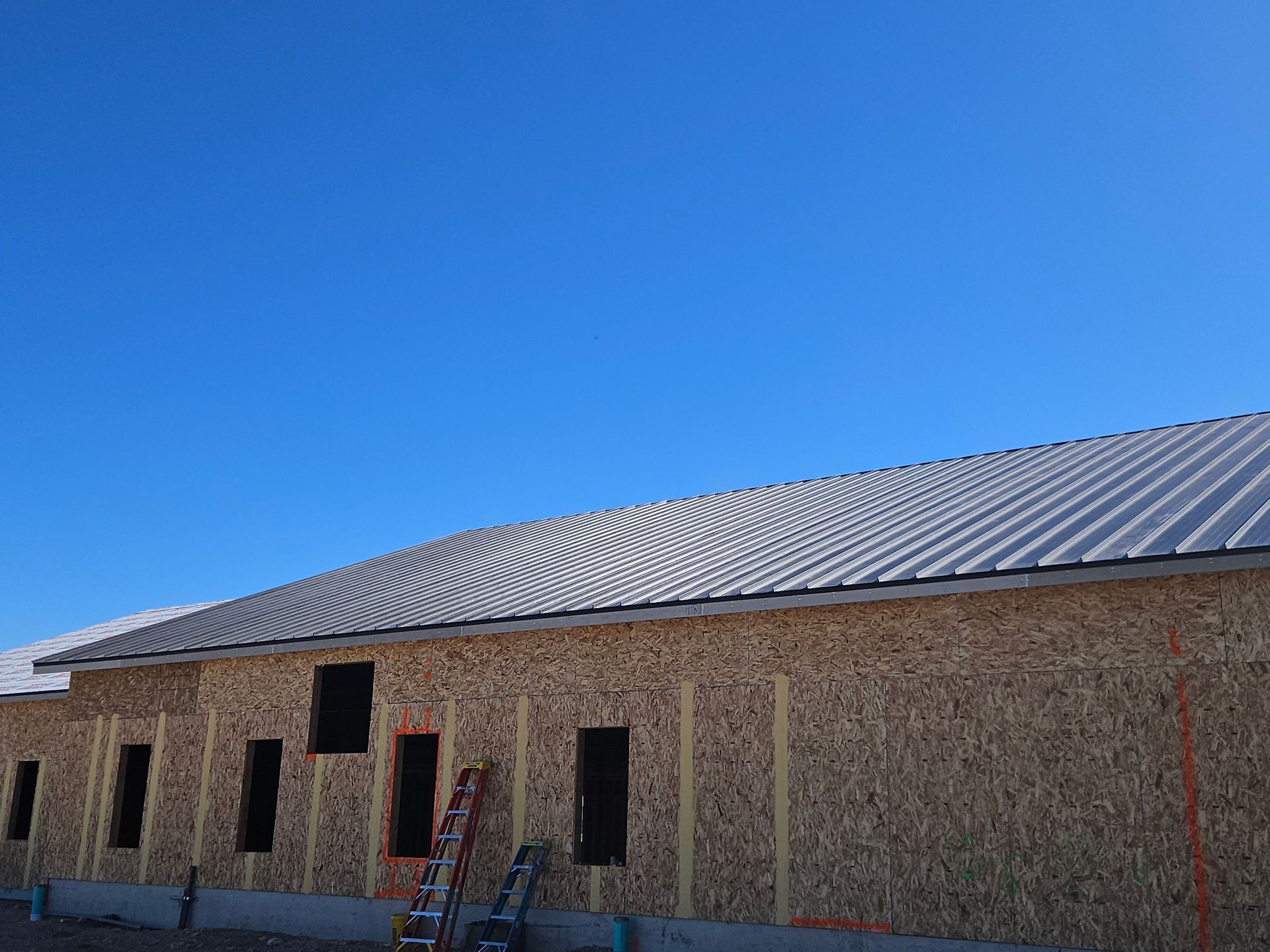 A building under construction with exposed OSB exterior walls, a metal roof, and a ladder against the wall under a blue sky.
