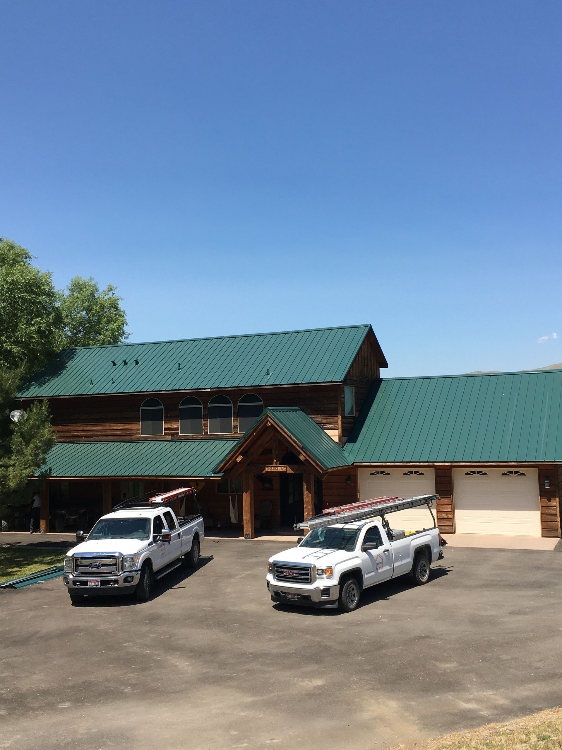 Two white work trucks with roof ladders parked in front of a log cabin home with a green metal roof under a clear blue sky.