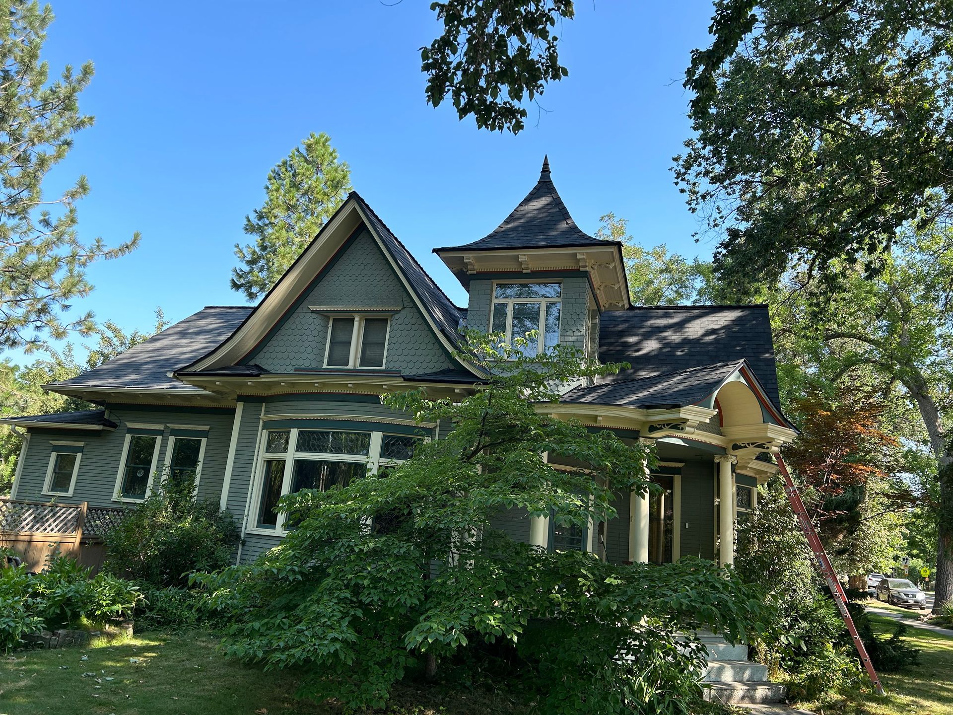 A Victorian-style two-story gray house with a peaked roof and a turret, partially obscured by a green tree.