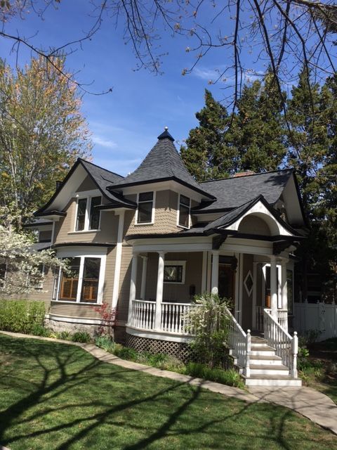 A tan, Victorian-style house with a wraparound porch, a central turret, and a dark roof, set on a sunny, grassy lawn.
