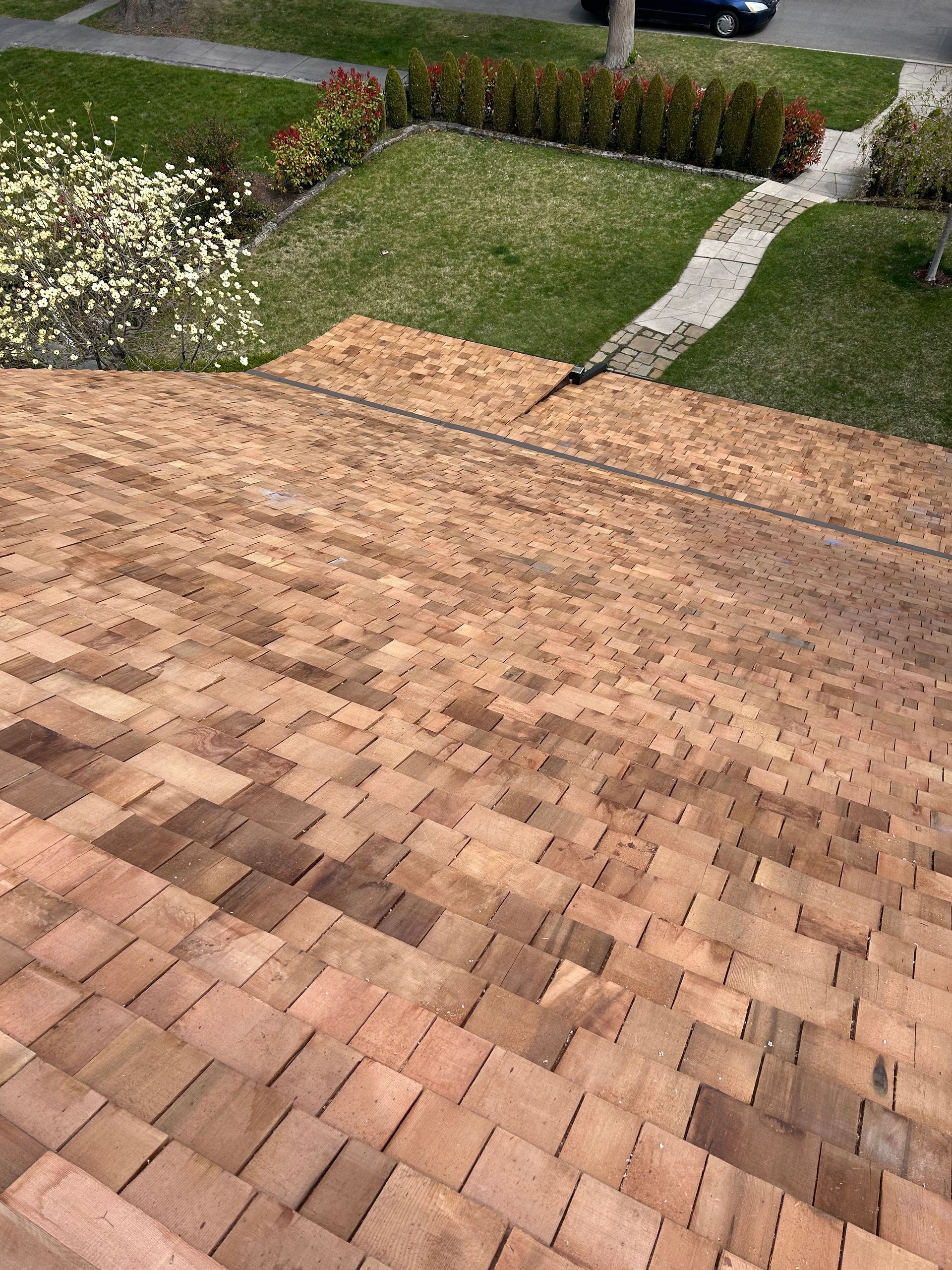A high-angle view looking down from a shingled roof onto a residential front yard with grass, a walkway, and trees.