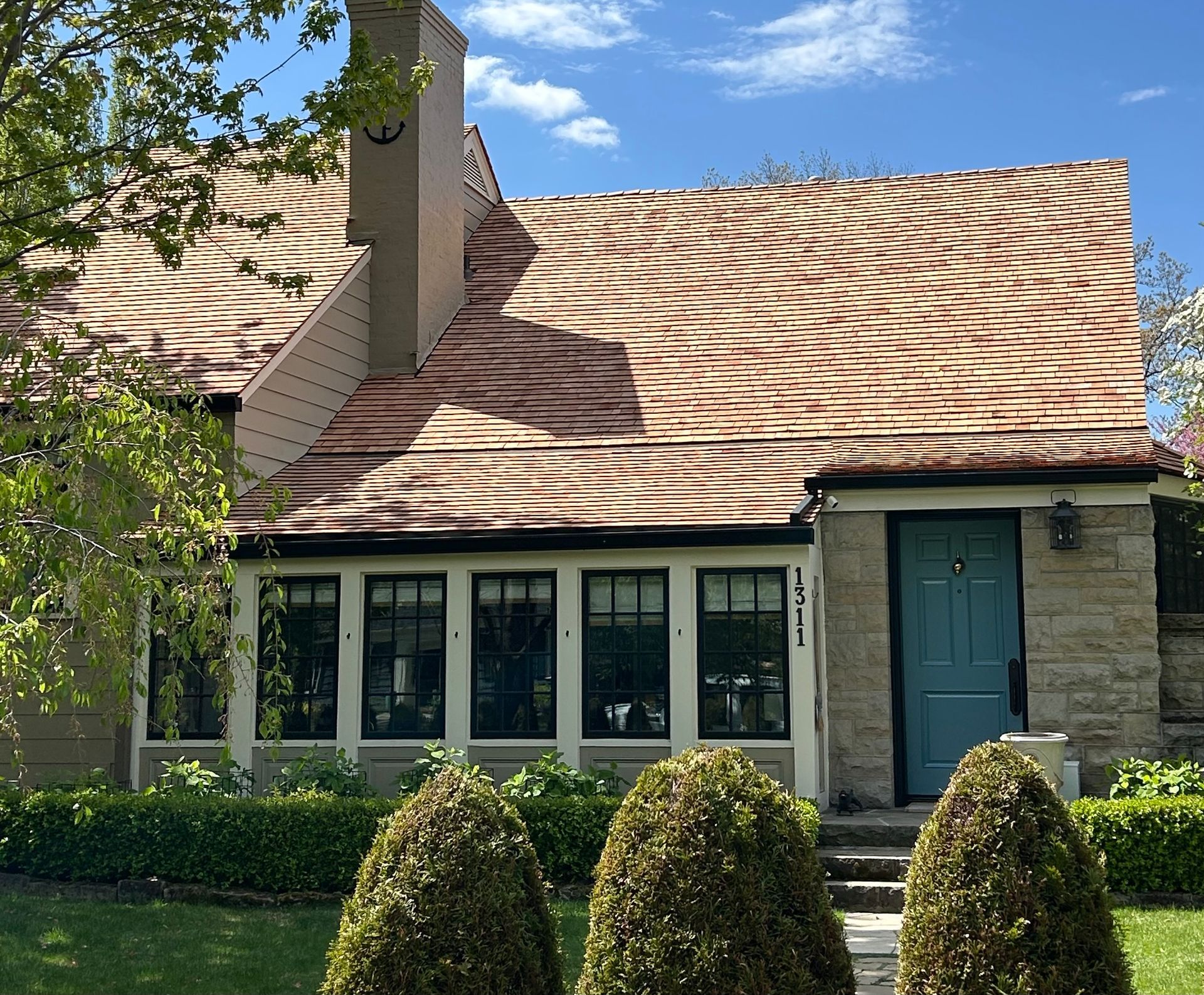 House with tan roof and blue door. Stone exterior, black-framed windows, manicured bushes, sunny day.