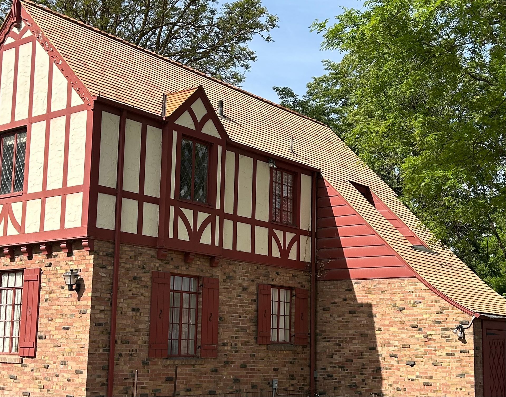 A Tudor-style building with red timber framing, light stucco, and stone walls, set against a green tree-filled background.