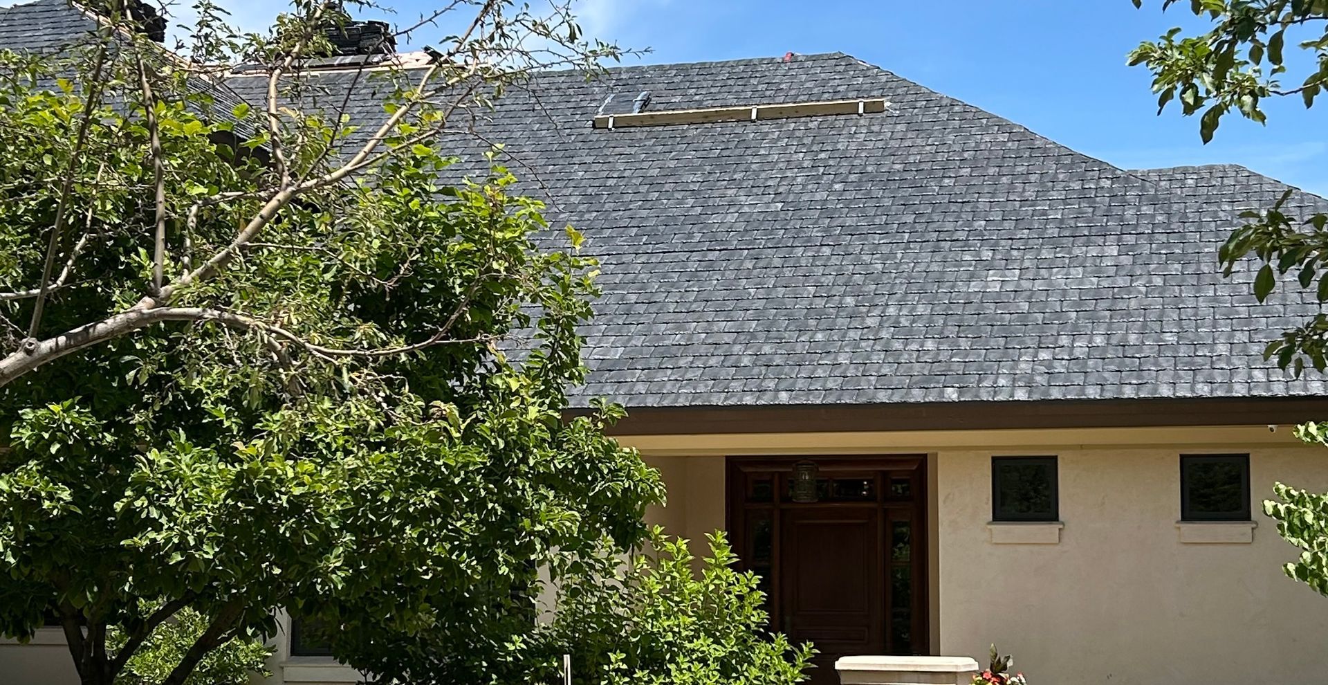 A house exterior with a grey shingled roof, a dark wooden front door, and light stucco walls, partially obscured by trees.