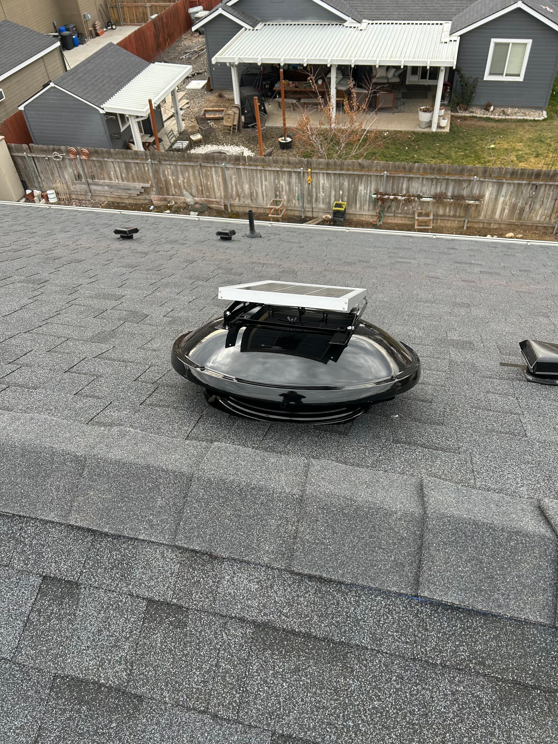 A solar-powered roof vent sits on a gray shingled roof, with a backyard and neighboring house visible in the background.