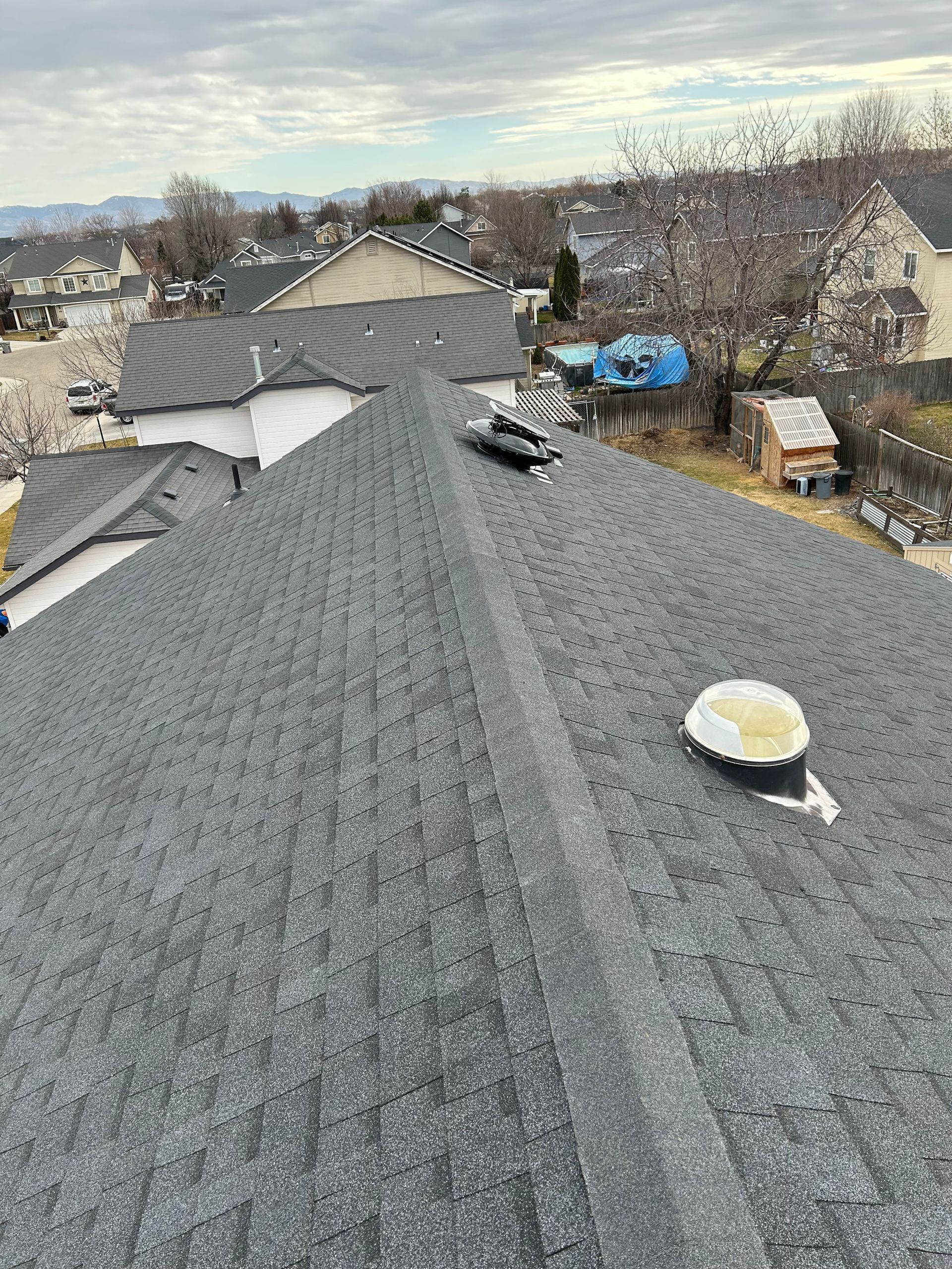 A high-angle view of a gray asphalt shingle roof with a prominent ridge, a vent pipe, and a piece of debris near the top.