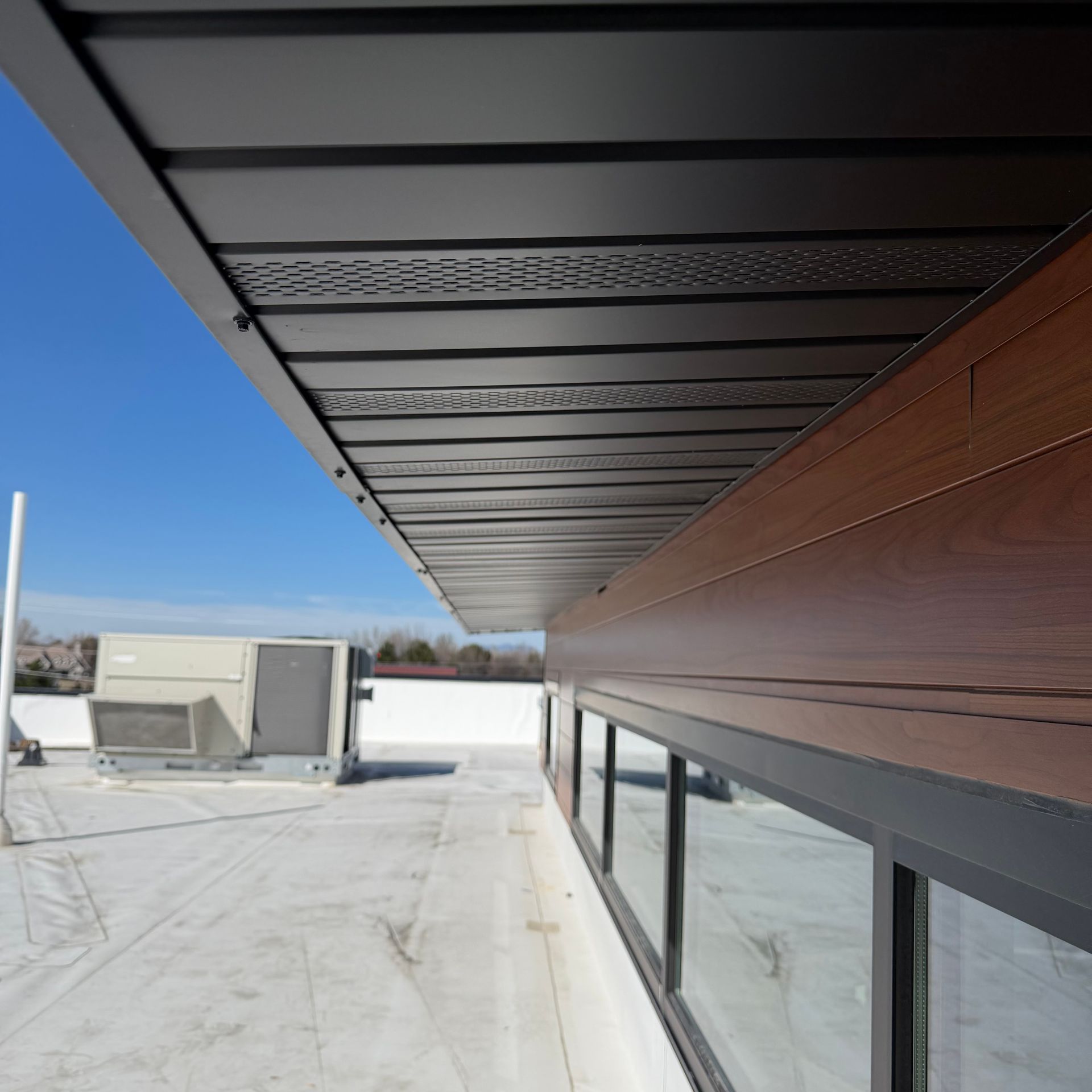 A low-angle view of a modern roof overhang with a wood-paneled wall, black metal trim, and an HVAC unit on a flat roof.