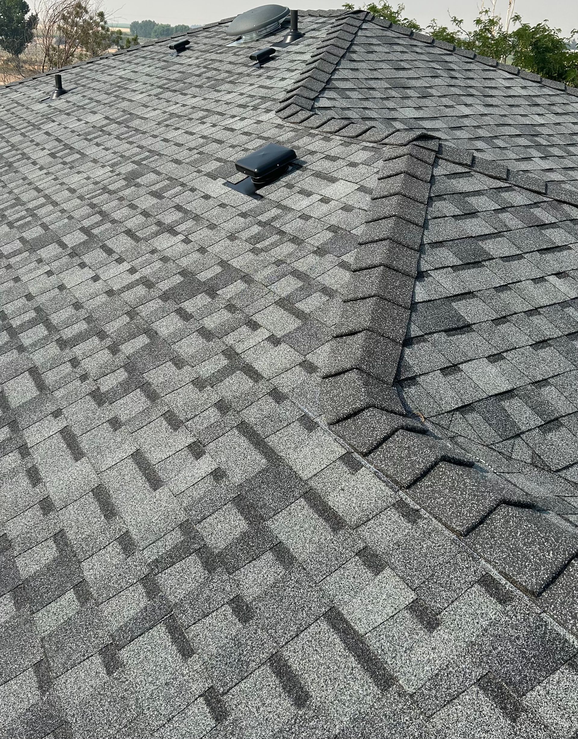 A high-angle view of a grey asphalt shingle roof featuring a hip and ridge vent, a vent pipe, and a circular skylight.