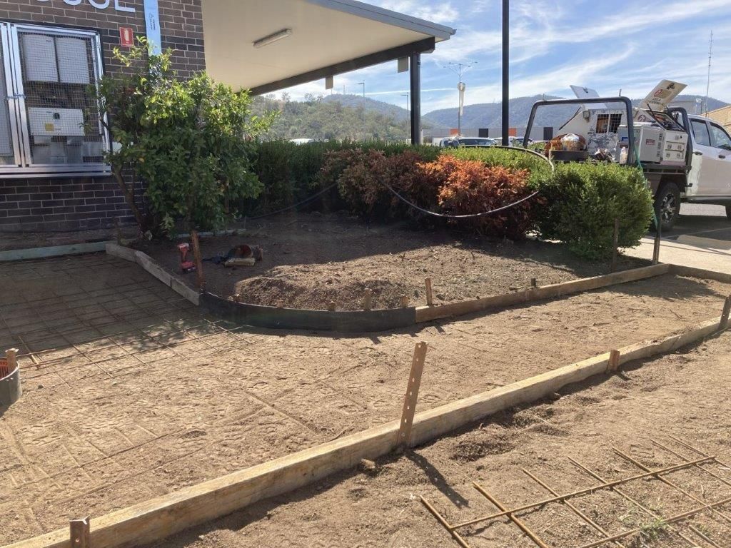 Construction site: wooden forms and rebar frame a pathway, in front of a building with landscaping.