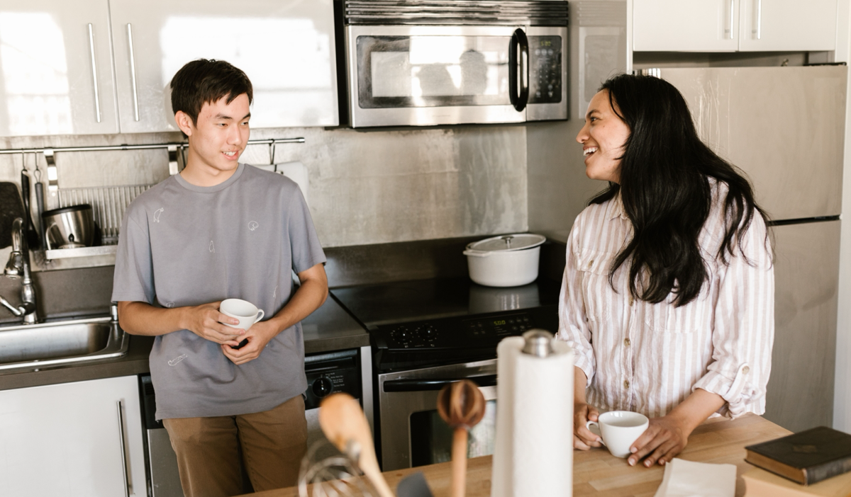 Two people smiling and talking in a bright kitchen, with a stove, microwave, and countertop items nearby