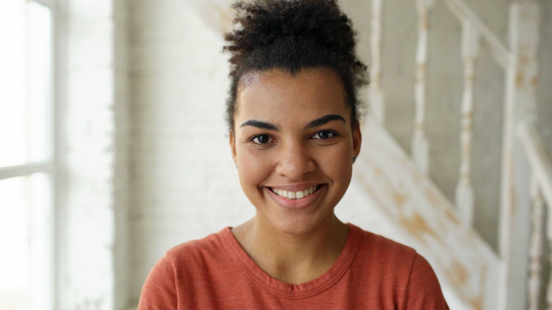Smiling person in an orange shirt standing indoors near a staircase.