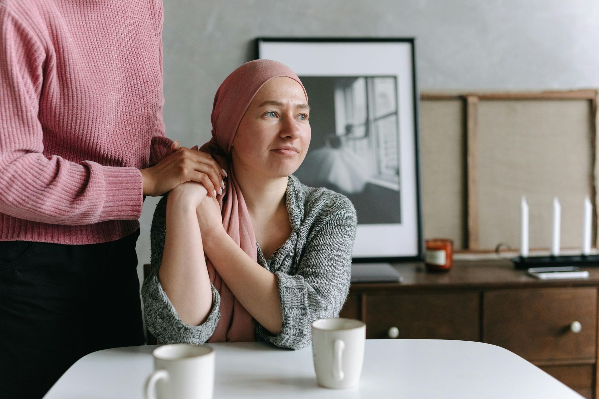 Person with a pink headscarf sits at a table while another person gently massages her shoulders in a cozy room.