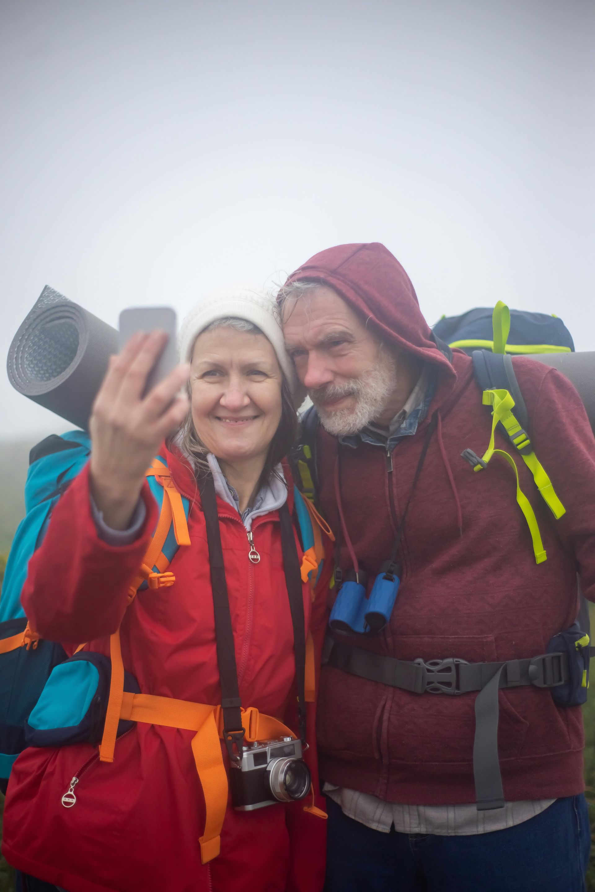An older man and woman with backpacks are taking a selfie on top of a mountain.
