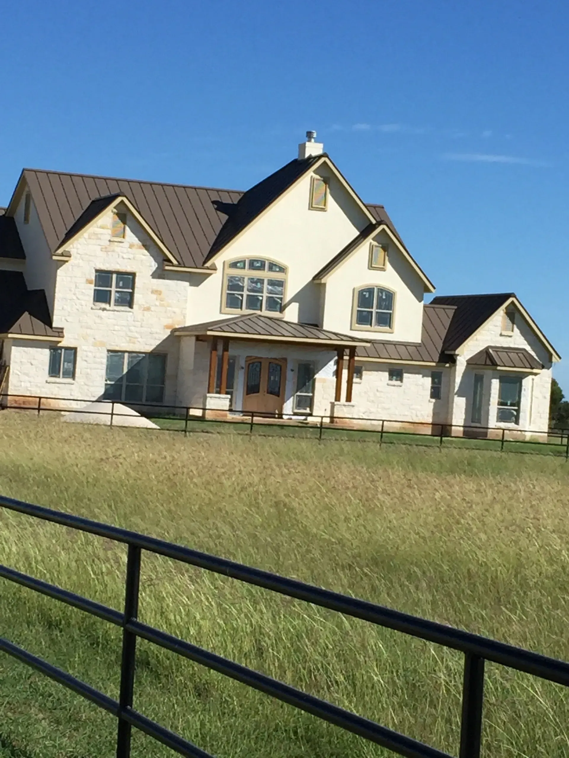 Two-story stone house with a brown roof and a porch. Tall grass and a black fence in the foreground. Blue sky.
