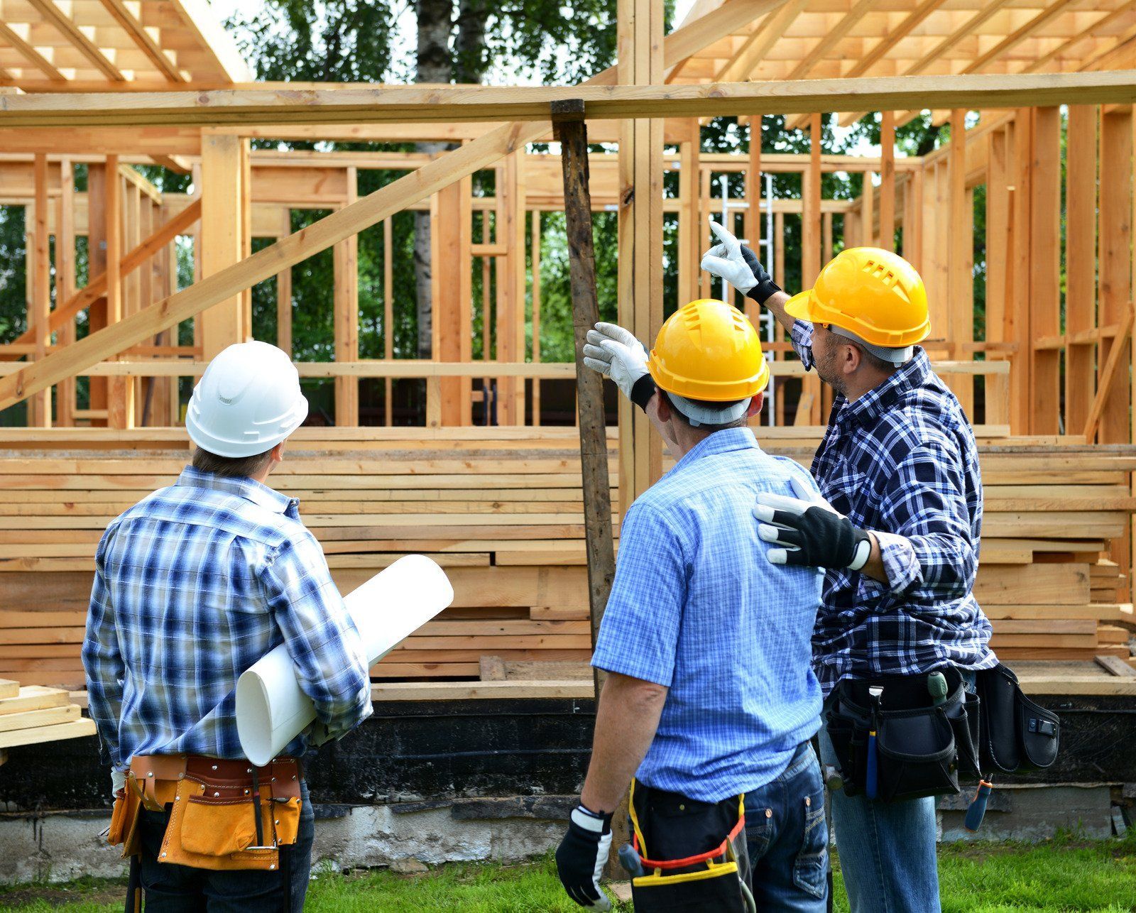 Construction workers examining a wooden house frame, holding plans and tools, wearing hard hats, outdoors.