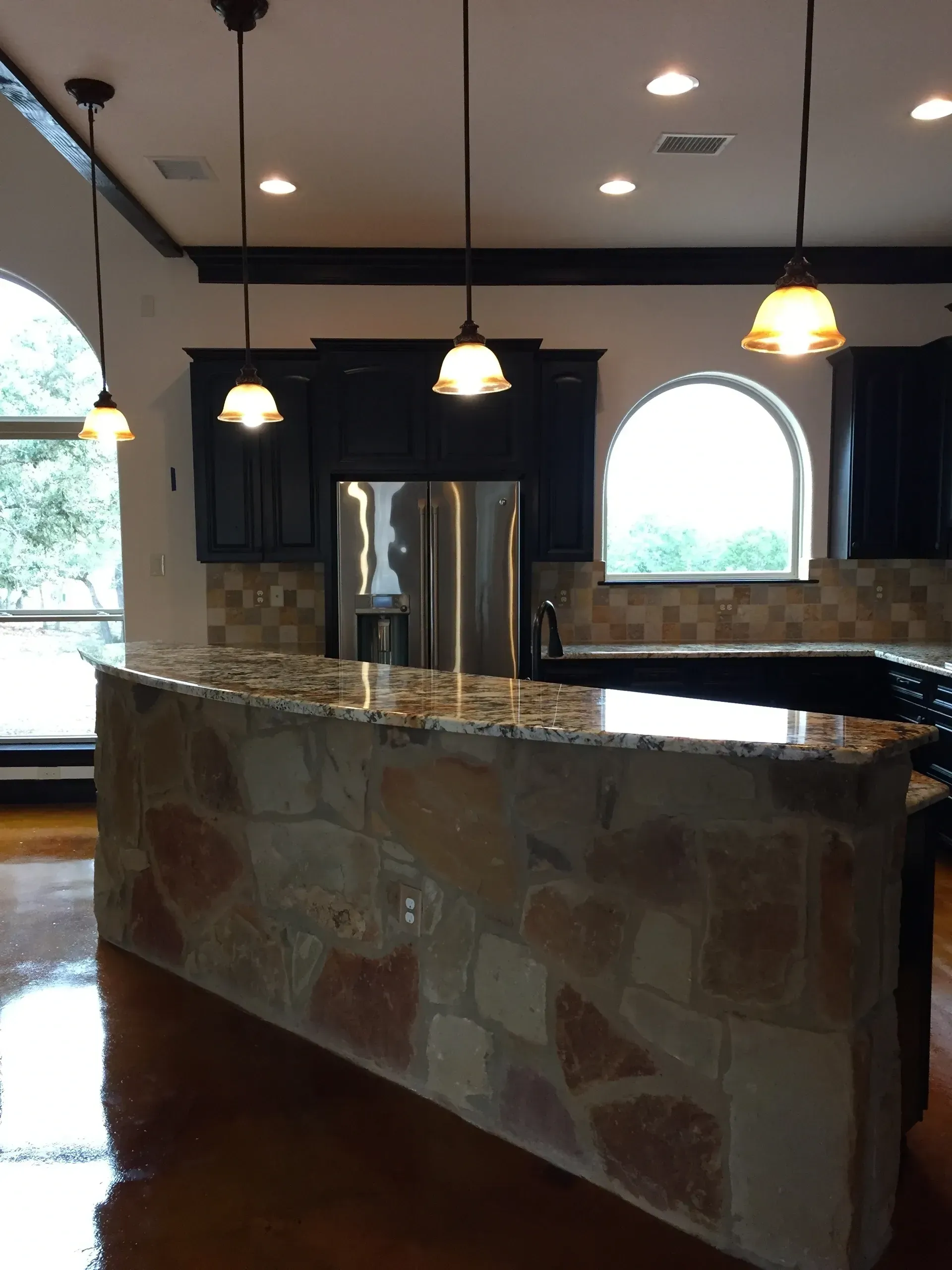 Kitchen with stone island, dark cabinets, granite countertop, pendant lights, and stainless steel refrigerator.
