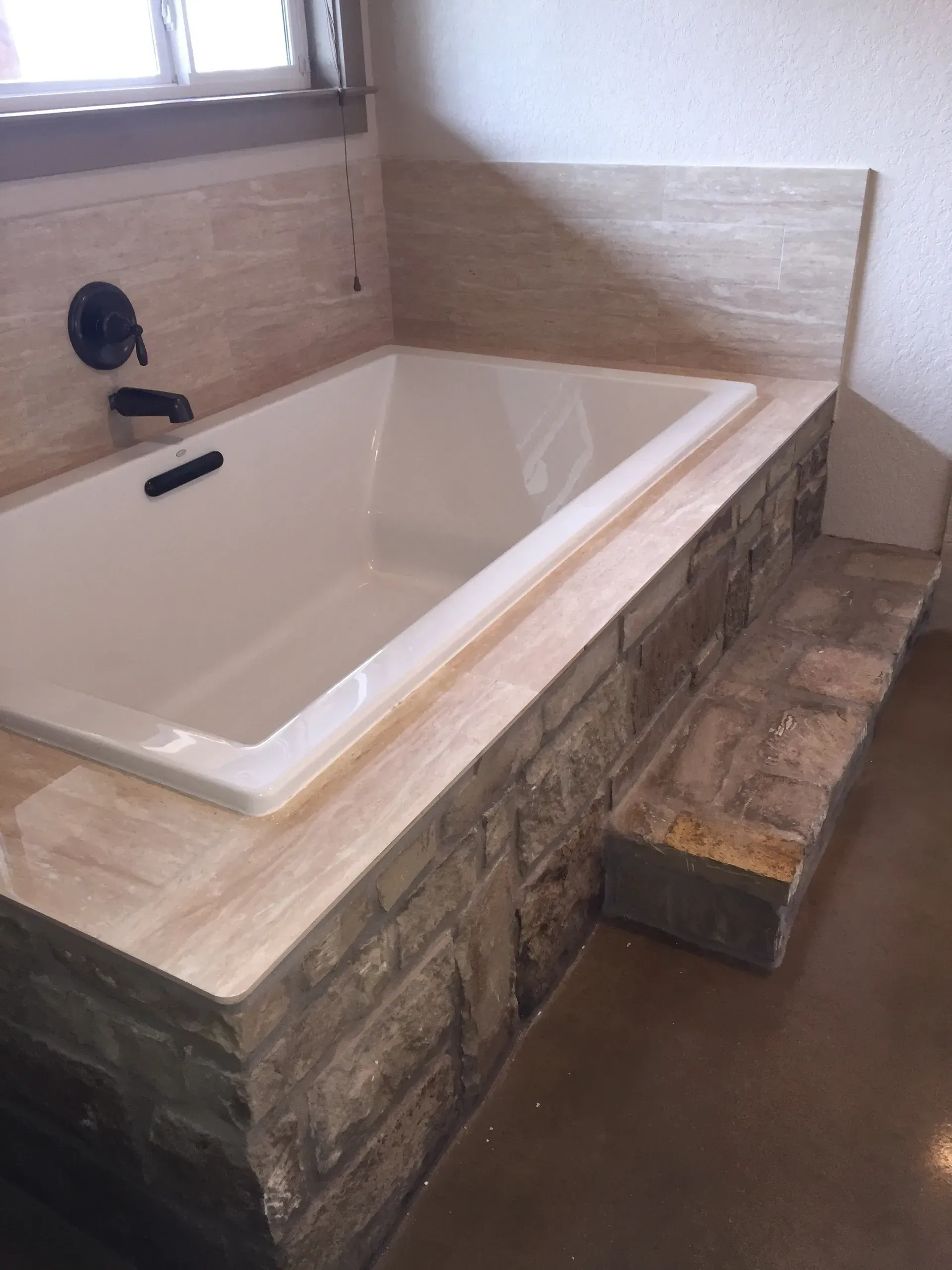 Built-in bathtub with stone surround and steps, next to a window. Black faucet. Light brown floor and walls.