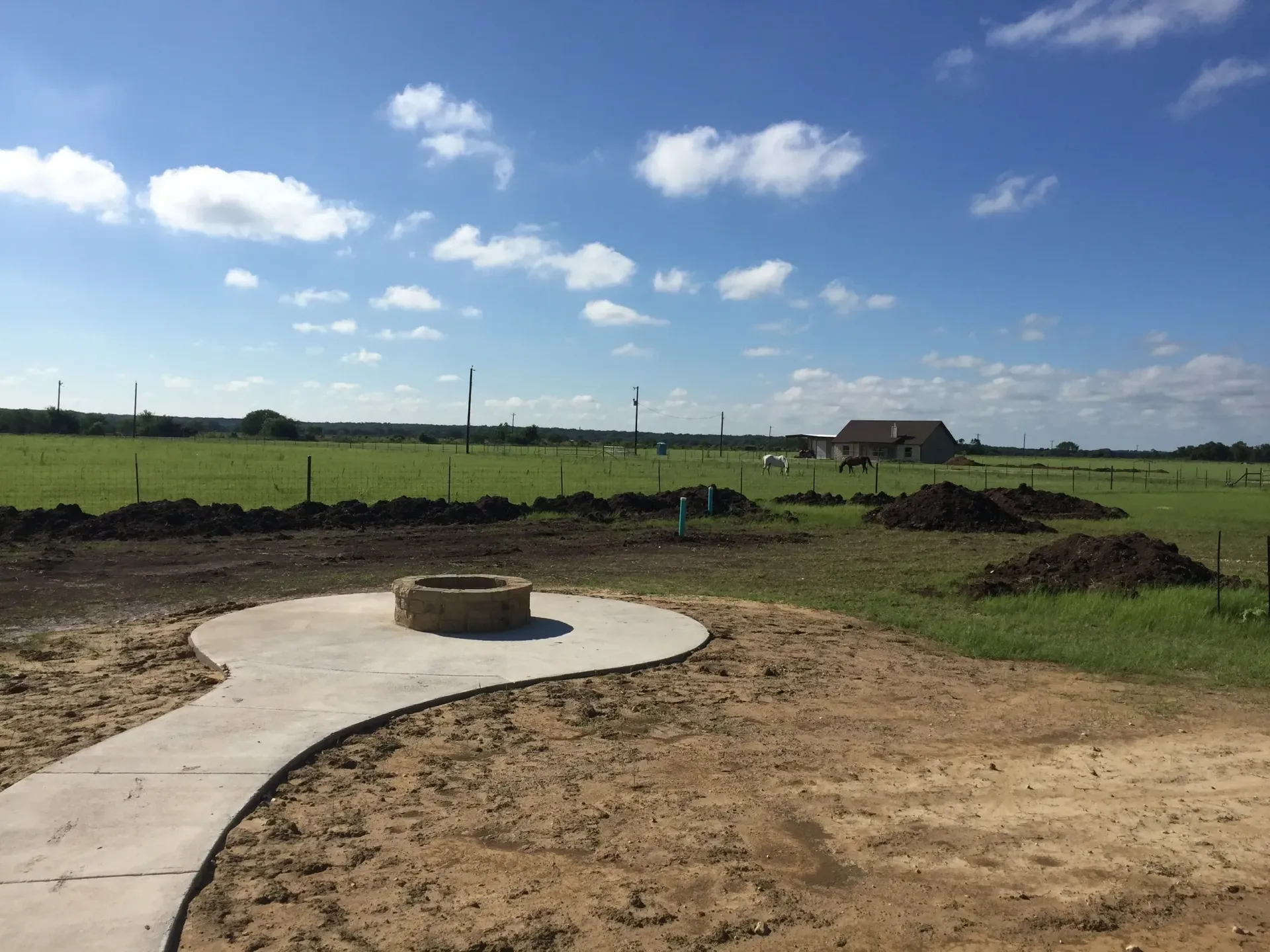 Concrete fire pit and pathway in a muddy field, with a house and blue sky in the background.