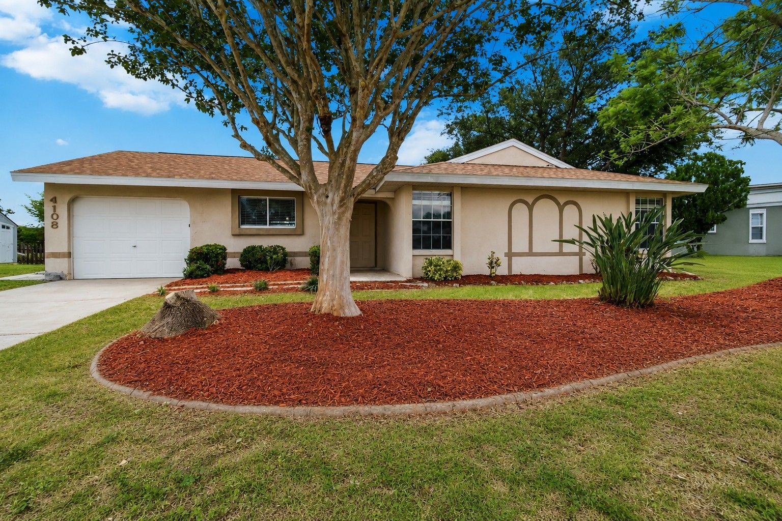 Single-story beige house with a white garage, large tree, and red mulch front yard