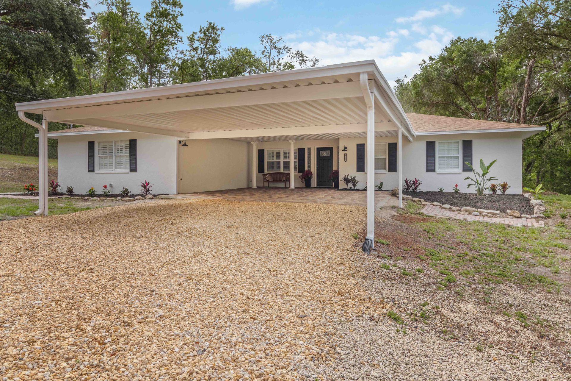 Single-story white house with a covered gravel driveway and carport, surrounded by trees