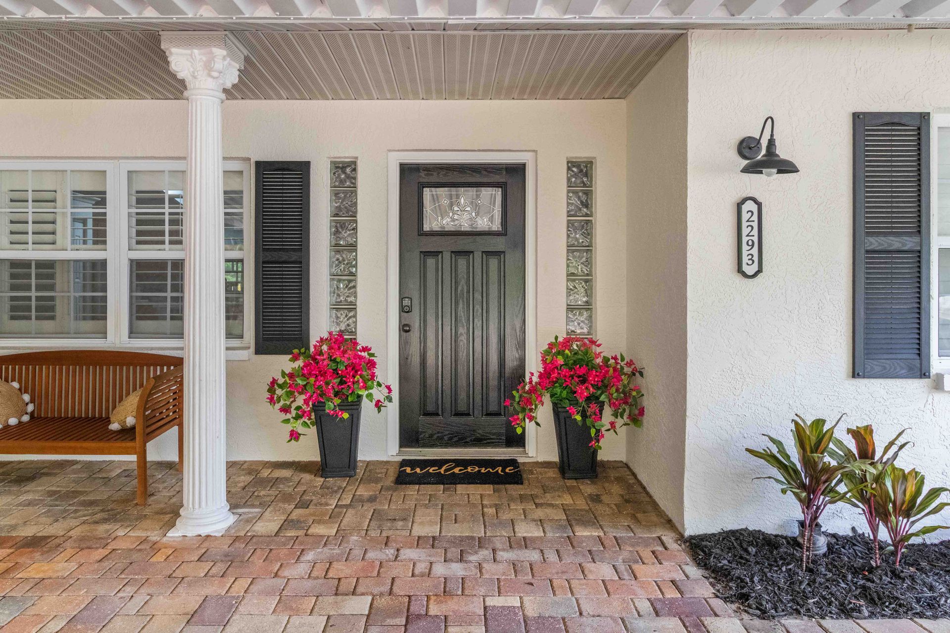 Front porch with black front door, white columns, brick pavers, and red potted flowers.